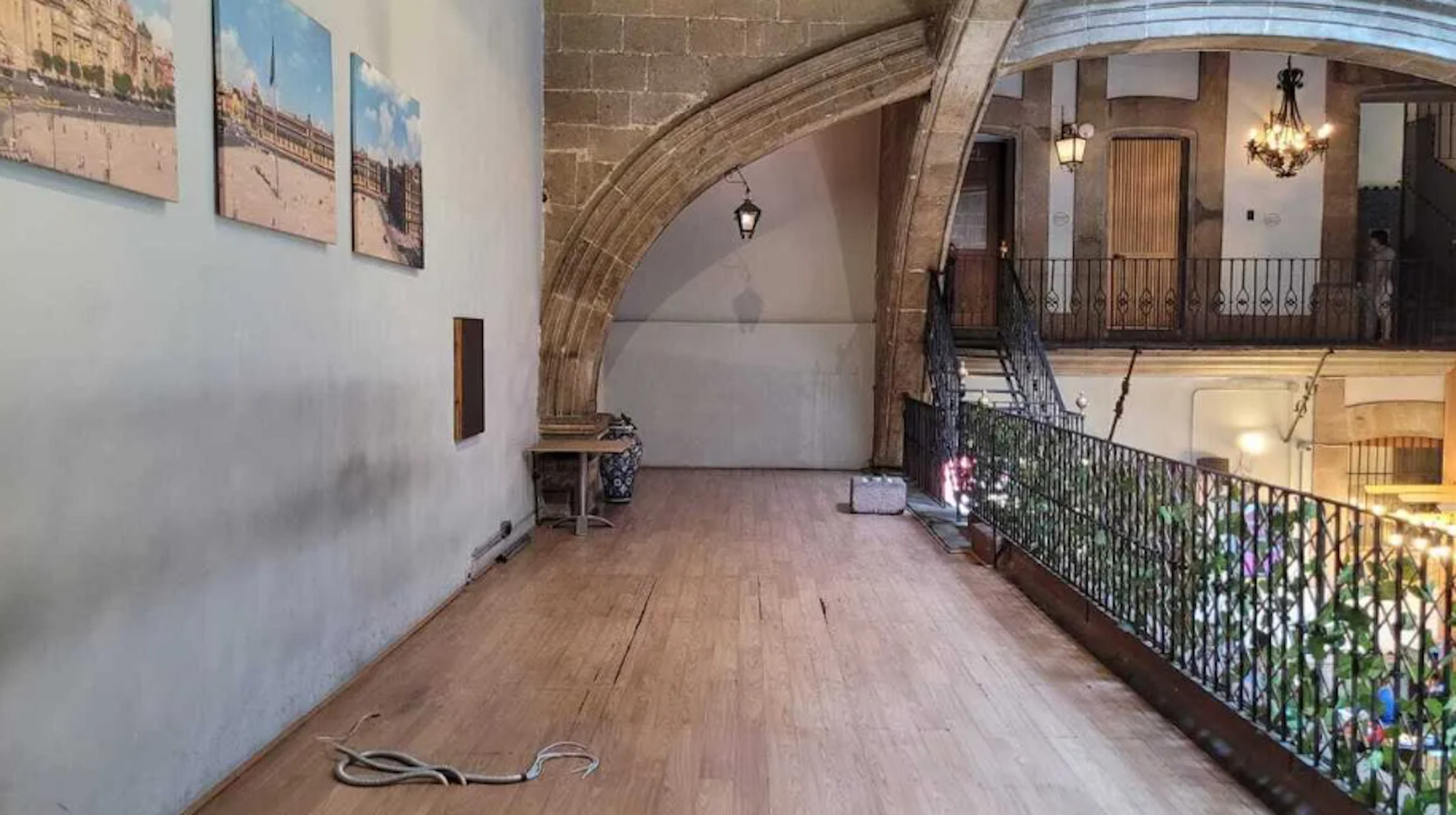 La imagen muestra un espacio interior elegante con arcos de piedra, suelo de madera y barandillas ornamentadas. Hay cuadros decorativos en la pared y un balcón con vistas a un nivel inferior. La arquitectura sugiere un edificio histórico renovado, posiblemente un hotel boutique o una residencia de lujo. El espacio combina elementos clásicos con toques modernos, creando una atmósfera acogedora y sofisticada.