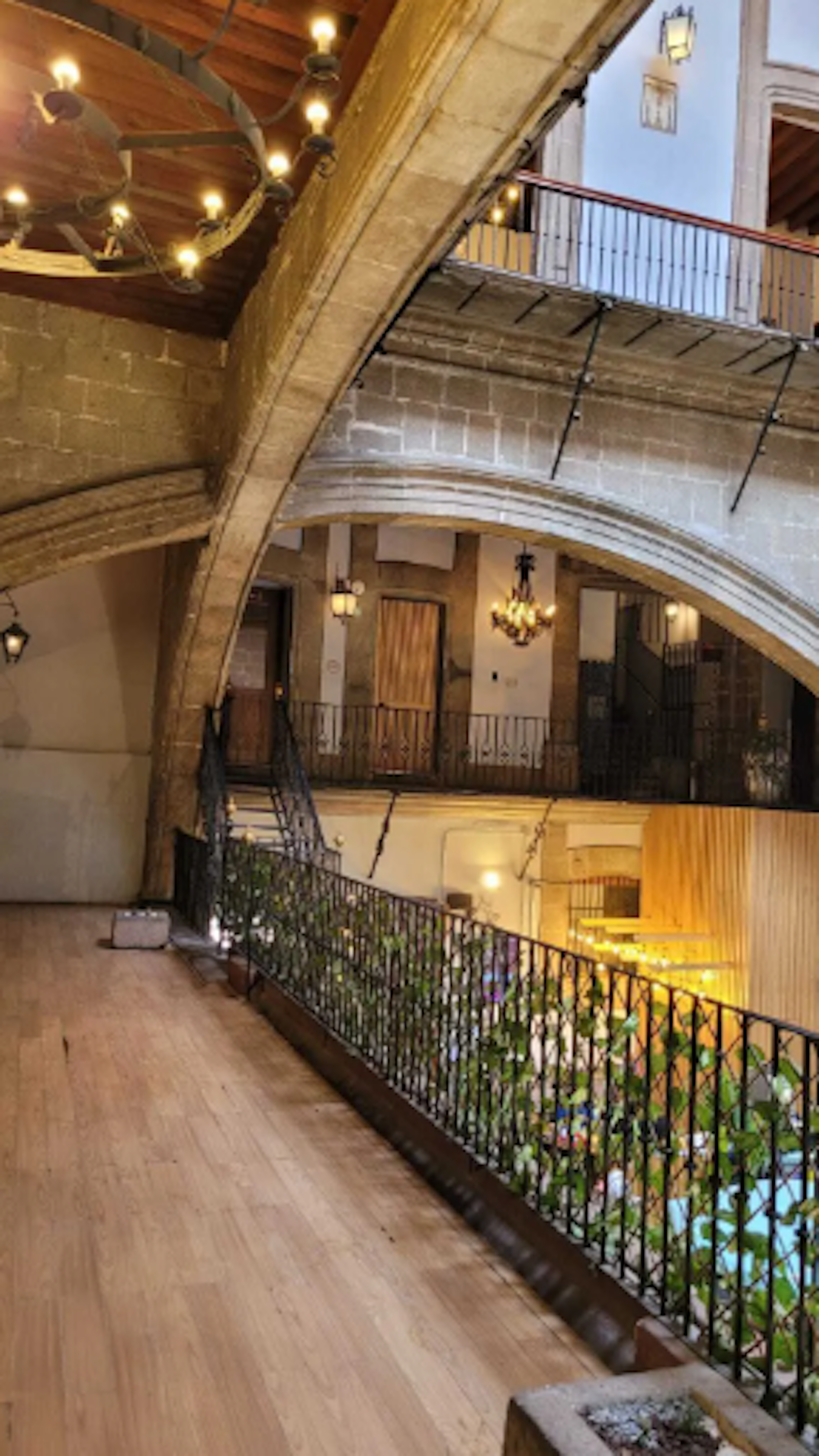 La imagen muestra el interior de un elegante edificio con arquitectura histórica, posiblemente un hotel boutique o una mansión convertida. Se aprecian arcos de piedra, balcones con barandillas de hierro forjado, pisos de madera y una iluminación cálida. El espacio combina elementos rústicos y lujosos, con plantas decorativas y una escalera que conduce a un nivel superior. El diseño sugiere un ambiente acogedor y refinado.