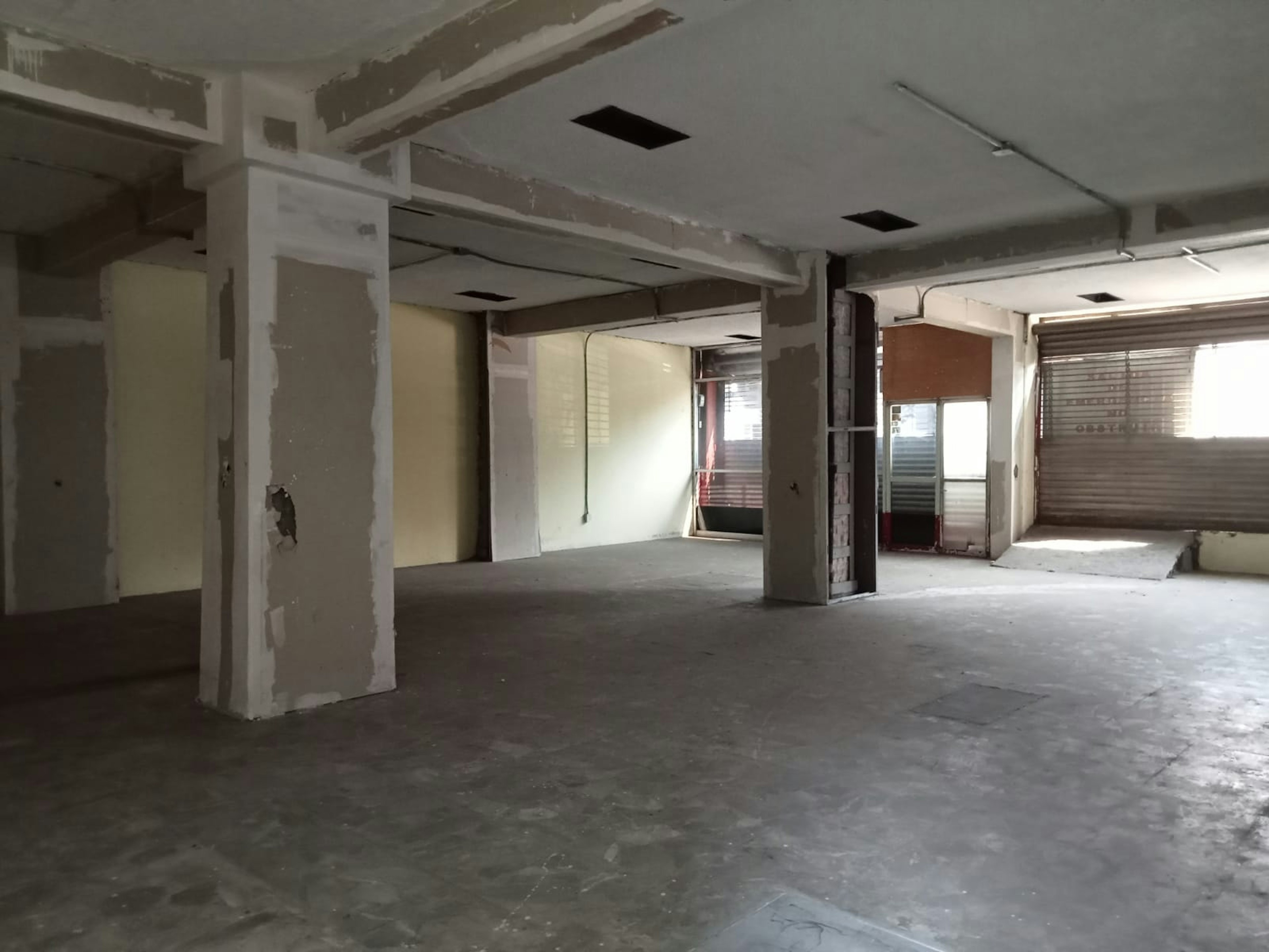 Esta imagen muestra un espacio interior en proceso de renovación. Se observa un área amplia y diáfana con columnas blancas, techos con vigas expuestas y suelo de cemento. Las paredes están parcialmente acabadas, con algunas áreas aún sin pintar. Hay ventanas con persianas en el fondo, sugiriendo que es un espacio comercial o industrial en transformación. El estado inacabado indica que está en plena fase de remodelación o construcción.