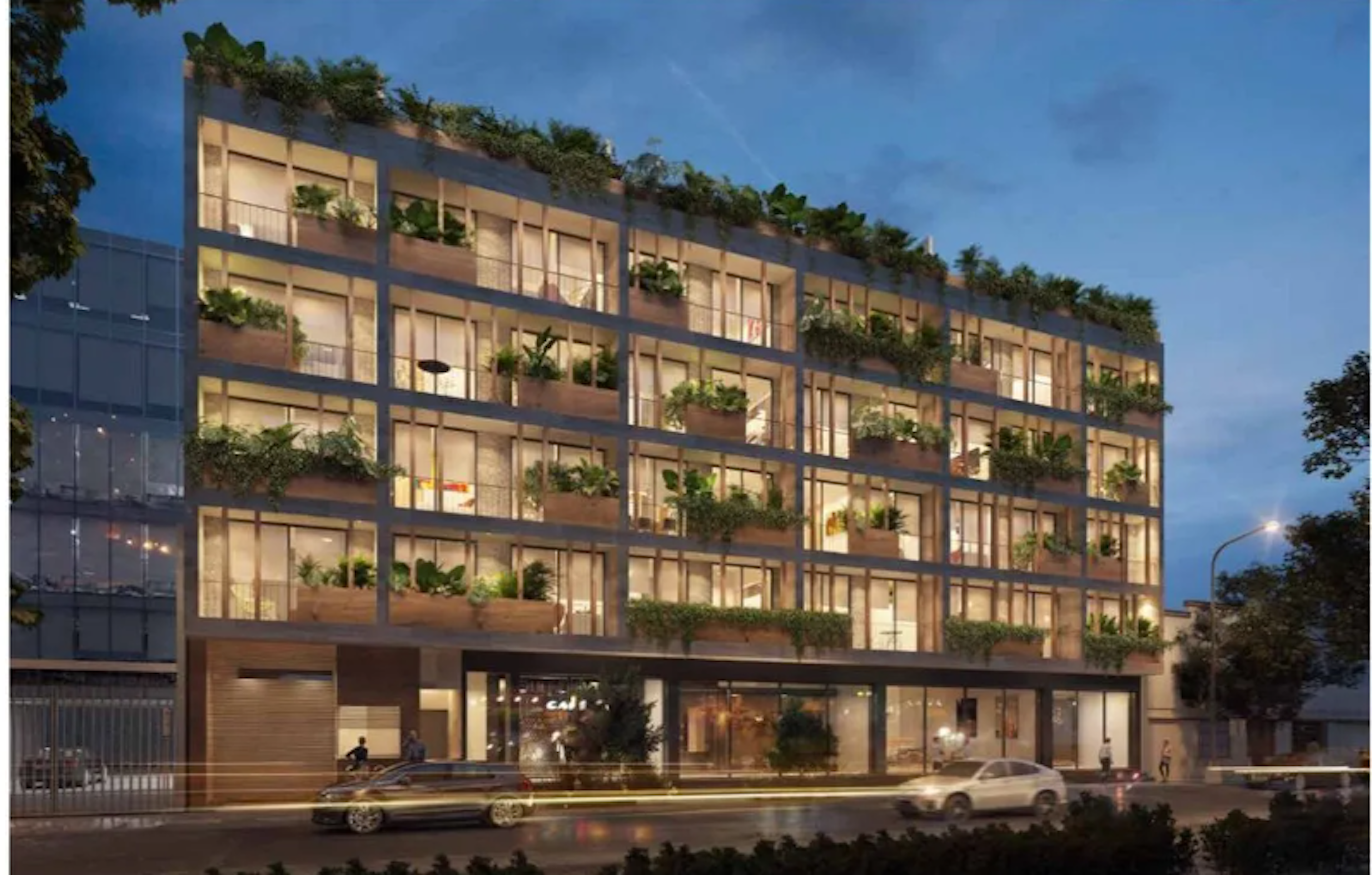 La imagen muestra un moderno edificio de apartamentos de varios pisos con un diseño ecológico. Cuenta con abundantes balcones y terrazas verdes, grandes ventanales iluminados y una fachada atractiva. En la planta baja hay locales comerciales. El edificio está iluminado en la noche, creando una atmósfera acogedora. Se observan algunos vehículos y personas en la calle, sugiriendo una ubicación urbana.