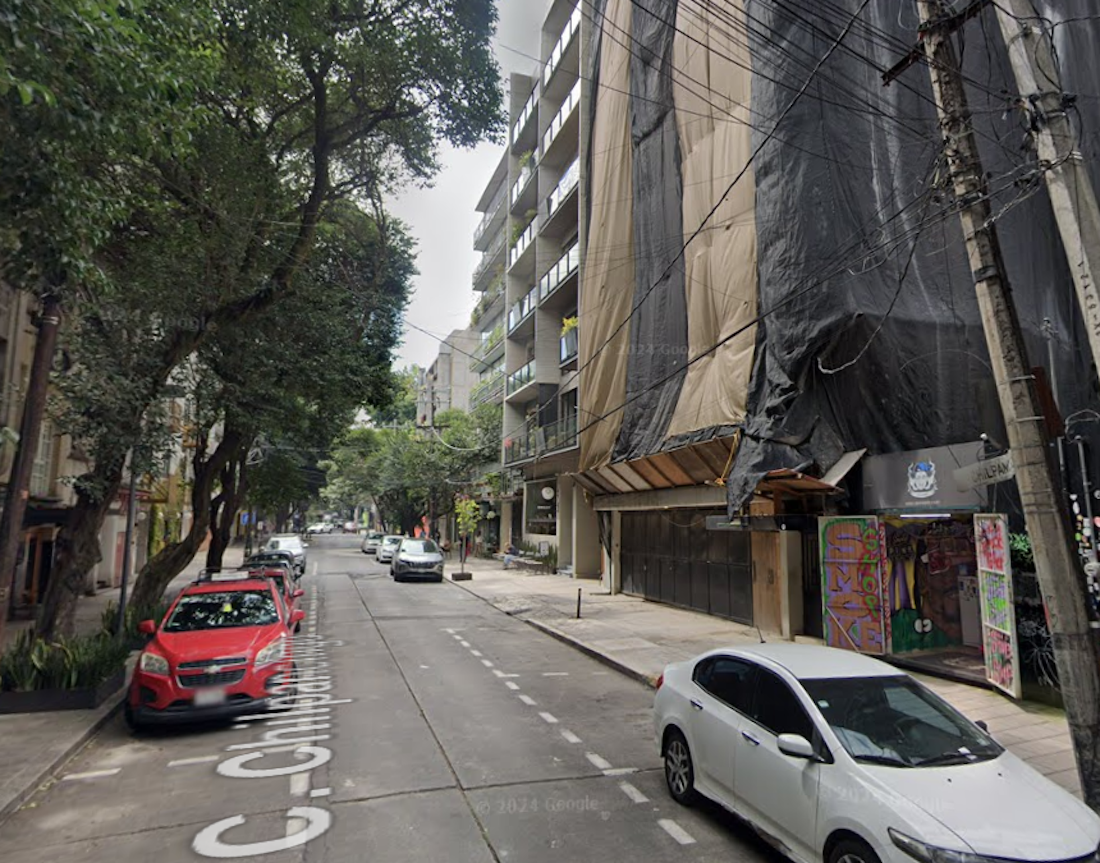 La imagen muestra una calle urbana con edificios residenciales de varios pisos. Se observa un edificio en construcción o remodelación cubierto con lonas. La calle está bordeada por árboles y cuenta con varios autos estacionados. Hay grafitis en algunas paredes, lo que sugiere un barrio con carácter artístico. La arquitectura mezcla estilos modernos y más antiguos, típico de una zona en desarrollo o gentrificación.