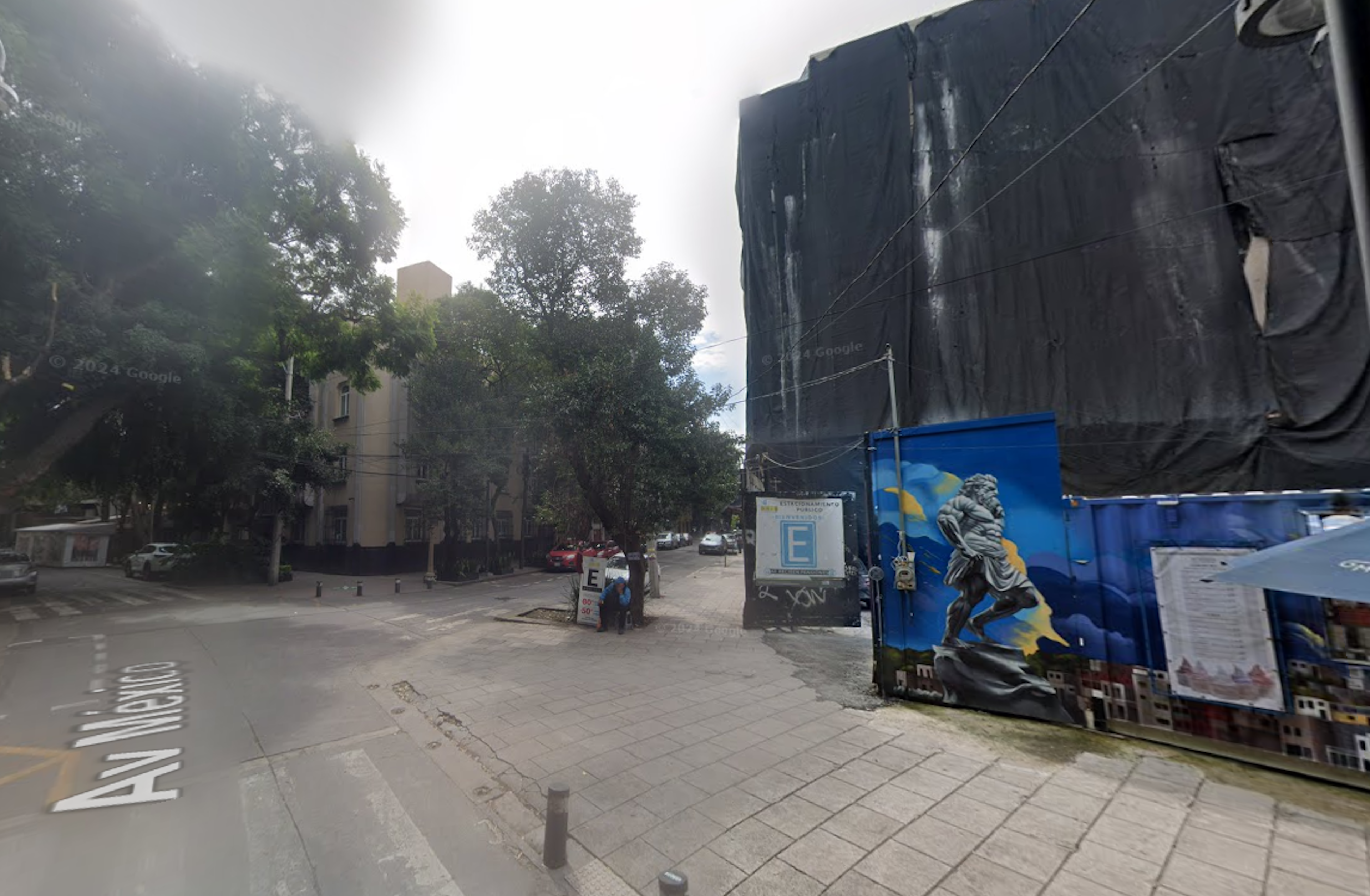 La imagen muestra una calle urbana con edificios residenciales y comerciales. Se observa una estructura cubierta con lona negra, posiblemente en construcción o renovación. Hay un mural colorido en una pared azul, árboles a lo largo de la calle, y señalización de estacionamiento. El pavimento es de adoquines y hay algunos vehículos estacionados. La escena sugiere un área en desarrollo o renovación urbana.