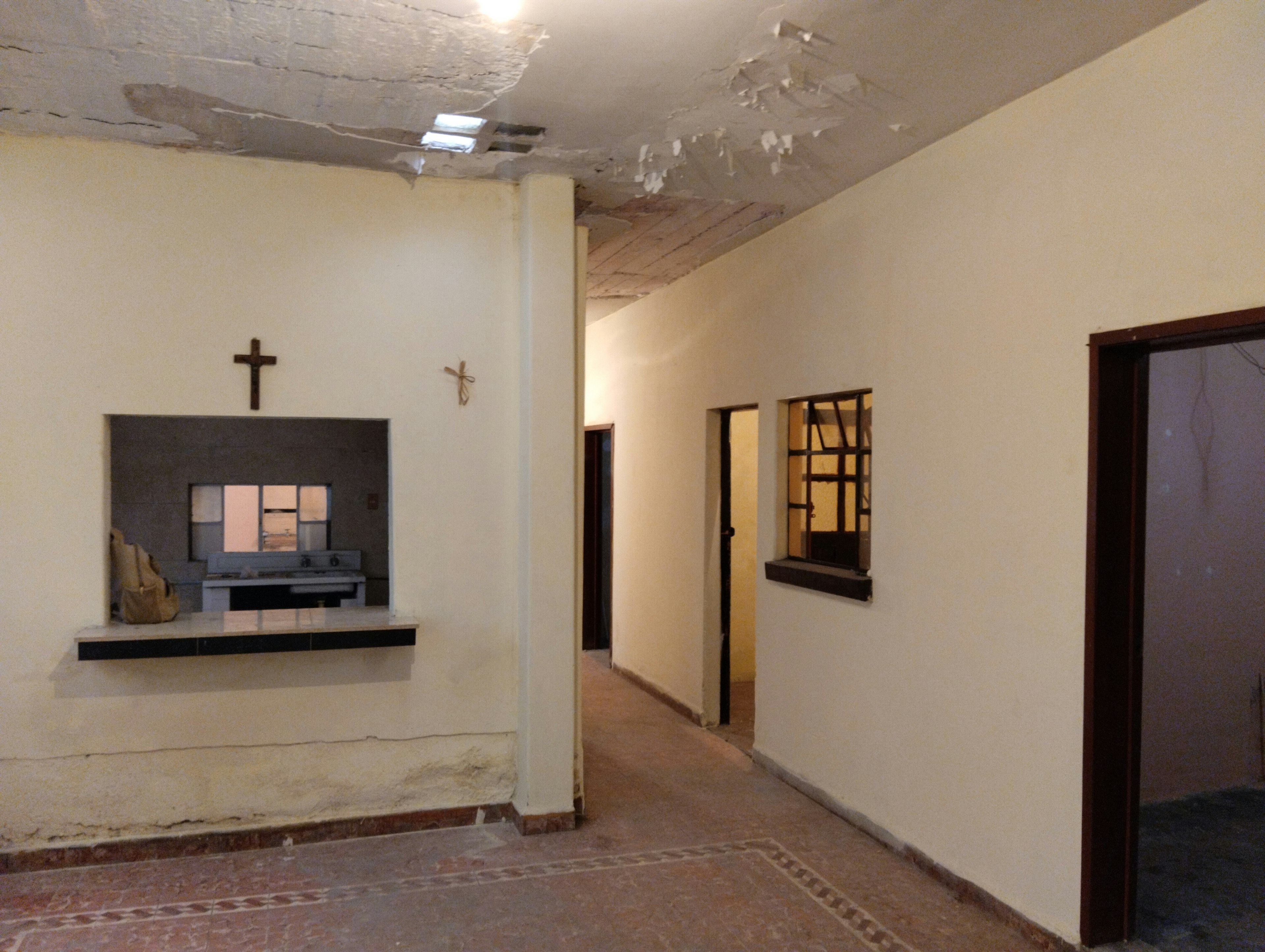 La imagen muestra el interior de una propiedad en proceso de renovación. Se observa un pasillo con paredes blancas, un techo dañado y un piso de baldosas. Hay una pequeña repisa con una cruz en la pared, sugiriendo un posible uso religioso previo. Se ven varias puertas y una ventana interior, indicando múltiples habitaciones. El estado general parece deteriorado, necesitando reparaciones significativas.