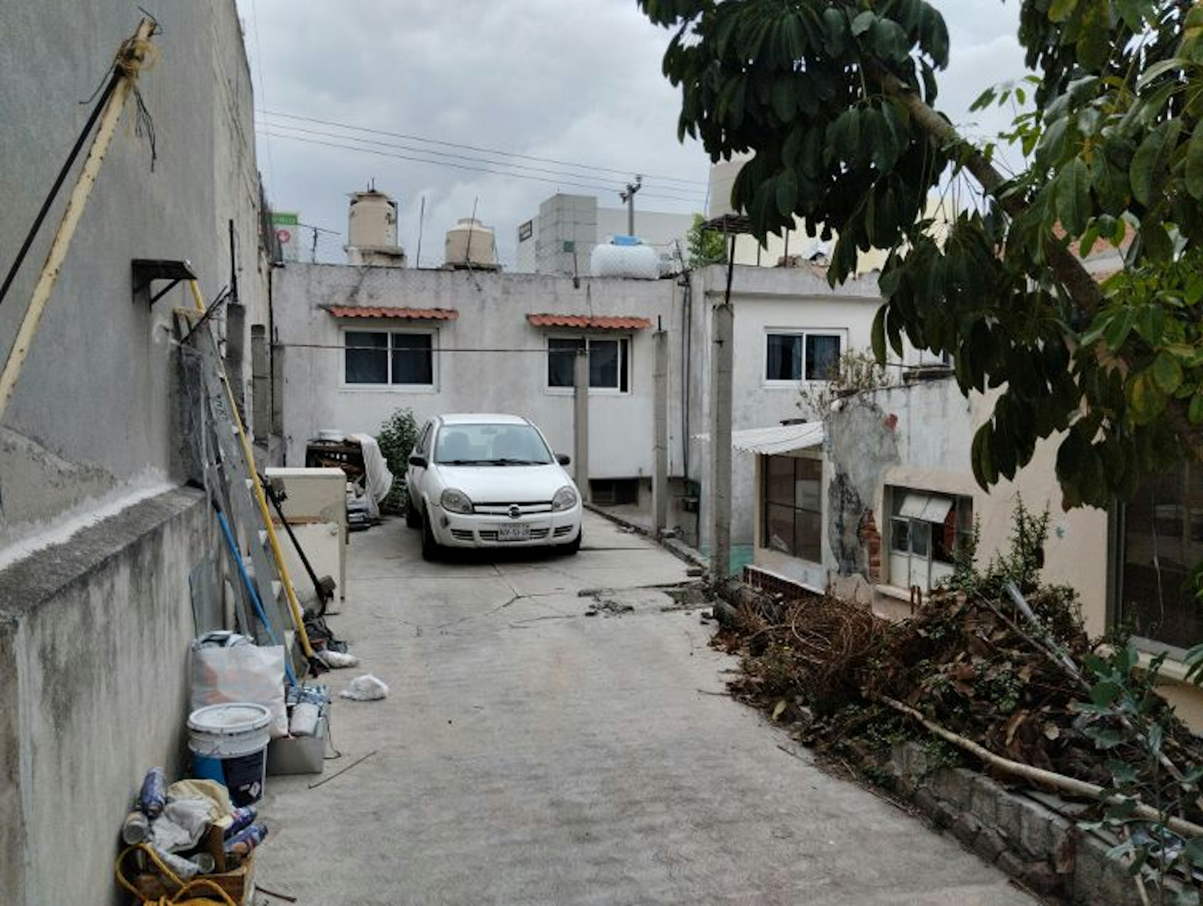 La imagen muestra un patio trasero de una propiedad residencial. Se ve una casa modesta con paredes blancas y ventanas pequeñas. Hay un auto blanco estacionado en el patio de concreto. El área tiene un aspecto descuidado, con escombros y vegetación dispersa. Se observan tanques de agua en el techo y cables eléctricos. El entorno sugiere una zona urbana de clase media-baja con potencial para mejoras y renovación.