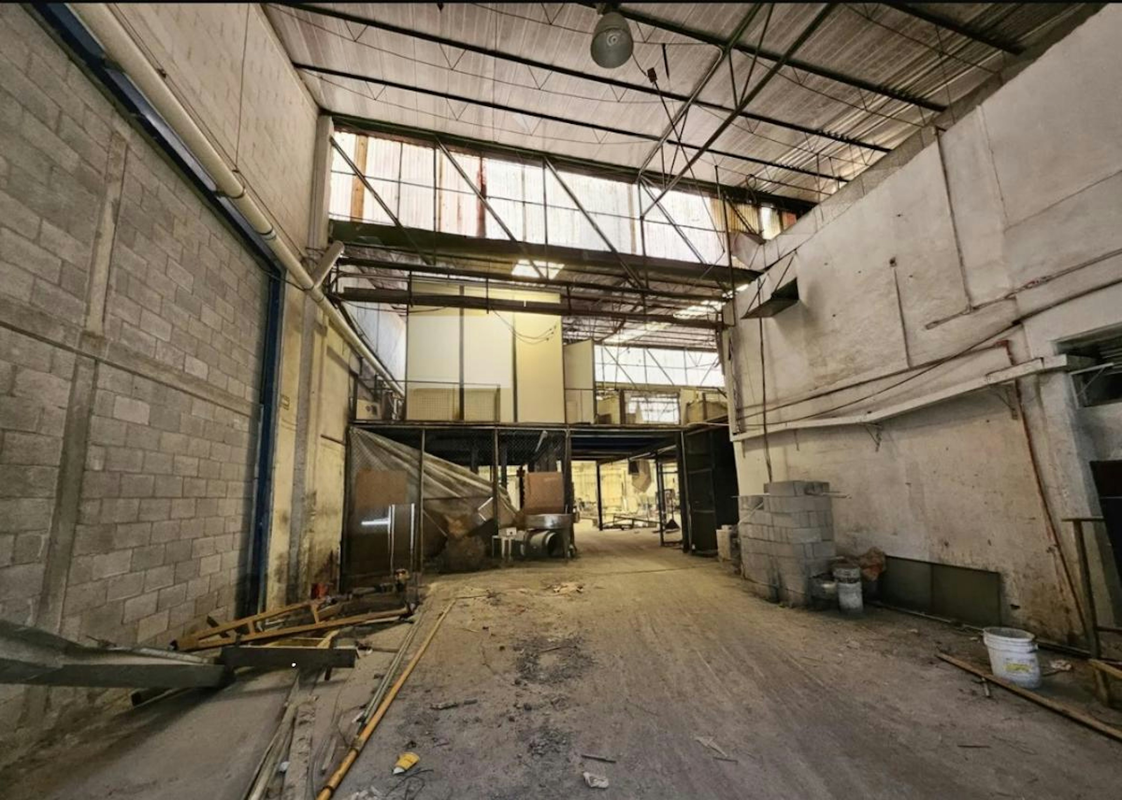 La imagen muestra el interior de un almacén o nave industrial en proceso de renovación. Se observan paredes de bloques, techos altos con estructura metálica y ventanas en la parte superior. El espacio está vacío y con escombros, indicando obras en curso. Hay un logo superpuesto que dice SIL Industry, sugiriendo que podría ser un proyecto de logística industrial. El estado inacabado sugiere potencial para personalización o remodelación.