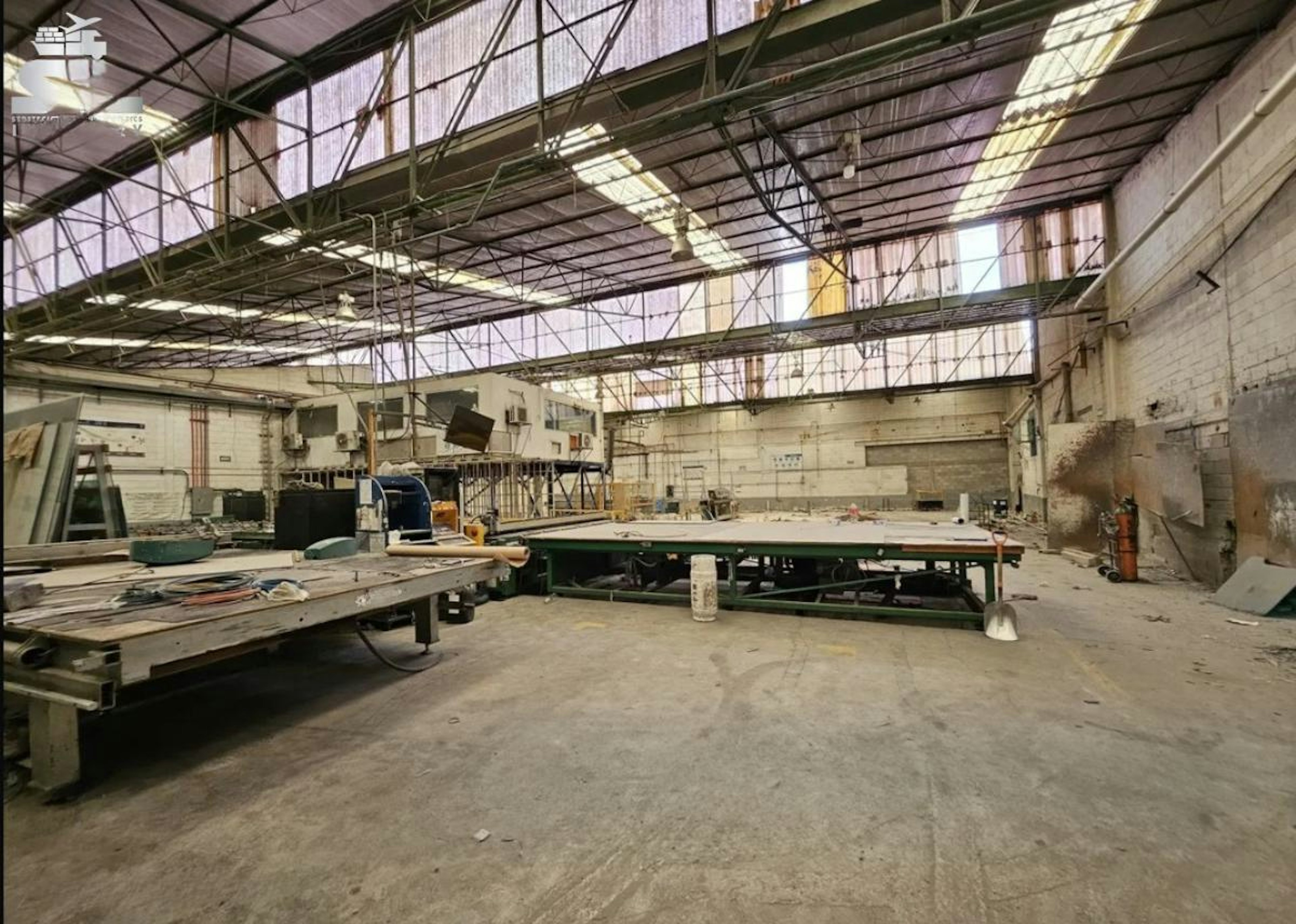 La imagen muestra un amplio espacio industrial con techo alto y estructura metálica. Se observan mesas de trabajo grandes, equipos y materiales dispersos, sugiriendo un área de producción o taller. El ambiente parece estar en proceso de remodelación o desuso parcial. Este tipo de espacio podría ser ideal para reconvertir en un loft industrial o un espacio comercial moderno, aprovechando su amplitud y carácter industrial.
