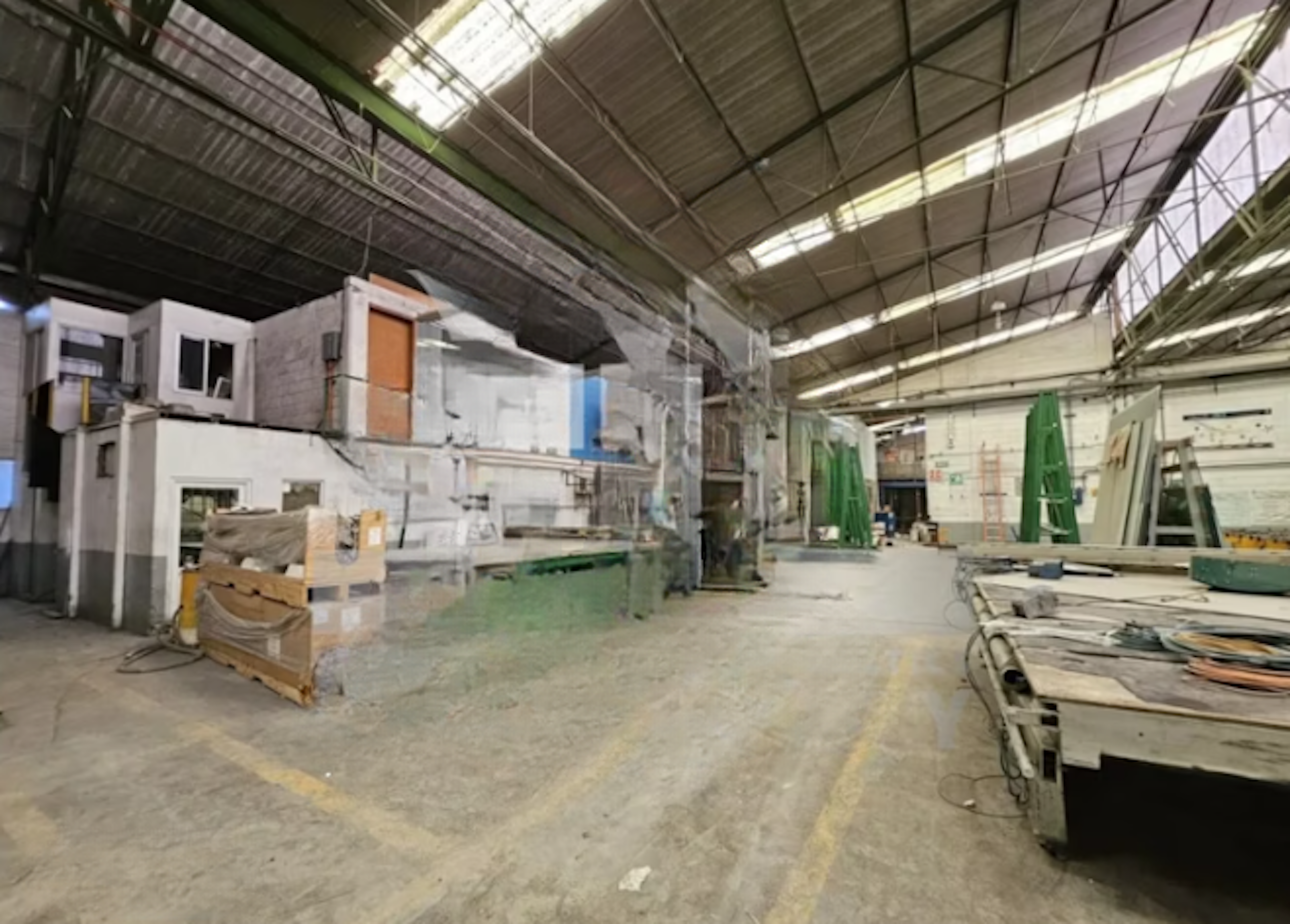La imagen muestra un amplio espacio industrial o de almacenamiento con techo alto y estructura metálica. Se observan paredes de ladrillo blanco, equipos y materiales de construcción dispersos. Hay áreas de trabajo, cajas y equipos almacenados. El espacio parece estar en proceso de remodelación o construcción, con zonas parcialmente terminadas y otras en obra. Este tipo de local podría adaptarse para diversos usos comerciales o industriales.