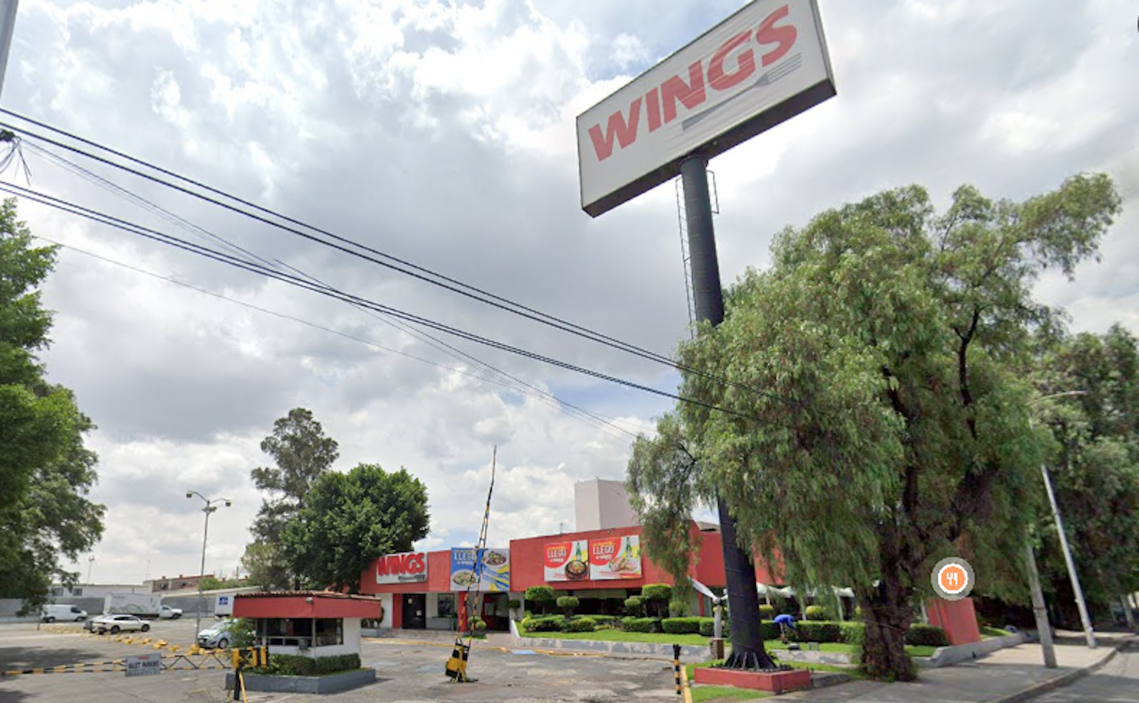 La imagen muestra un local comercial de la cadena de restaurantes Wings. Se observa un gran letrero publicitario con el nombre de la marca, un edificio rojo con anuncios y un estacionamiento. El entorno incluye árboles, postes eléctricos y una calle. El cielo está nublado, sugiriendo un día gris. La propiedad parece estar ubicada en una zona comercial con tráfico vehicular y peatonal.