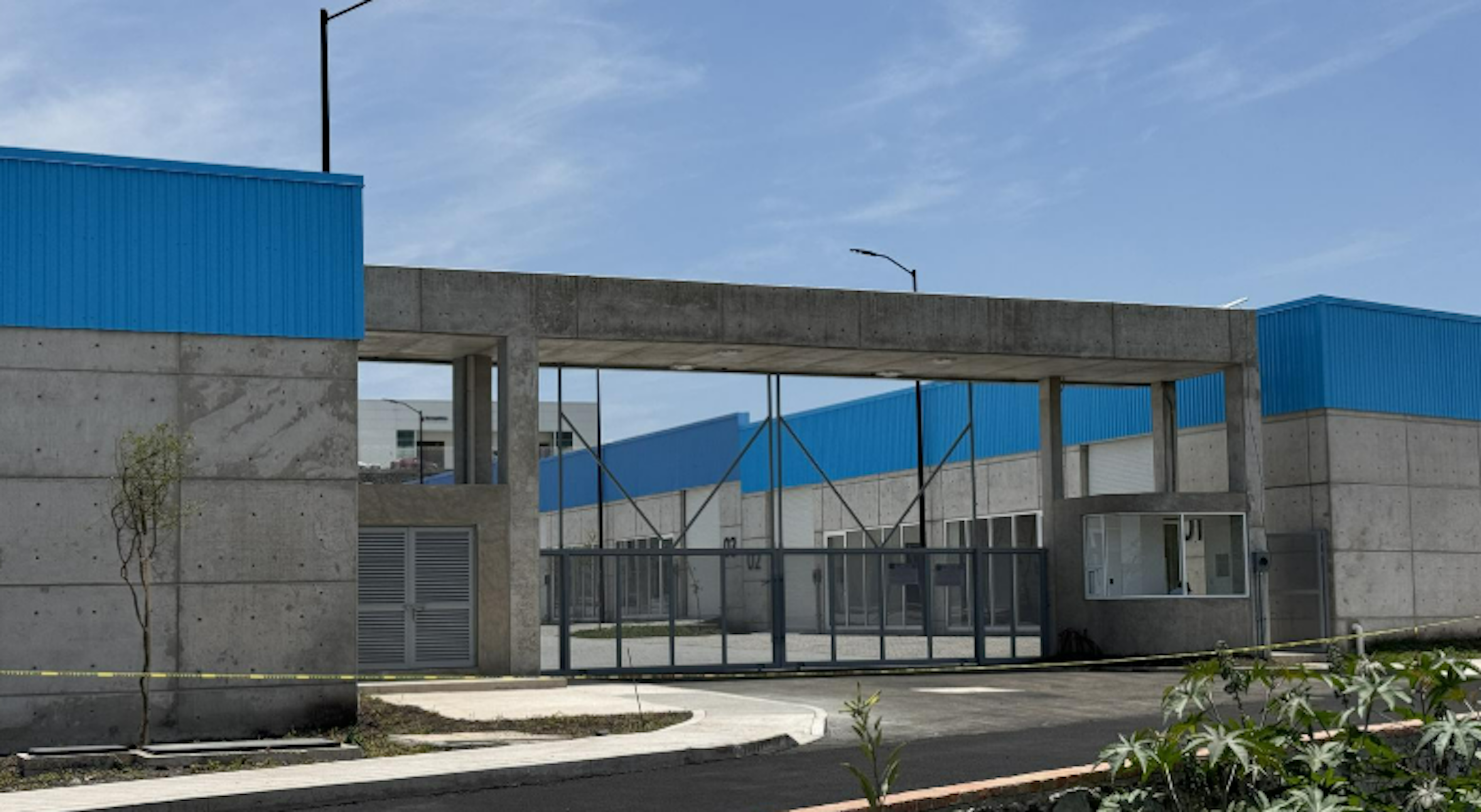 La imagen muestra la entrada de un complejo industrial o almacén. Se ve una estructura de concreto con un gran portón de metal azul. El edificio tiene paredes de concreto gris y techos azules. Hay una pequeña caseta de seguridad visible y algunas áreas verdes con plantas jóvenes. El cielo es azul claro, sugiriendo un día soleado. La arquitectura es moderna y funcional, típica de zonas industriales o comerciales.