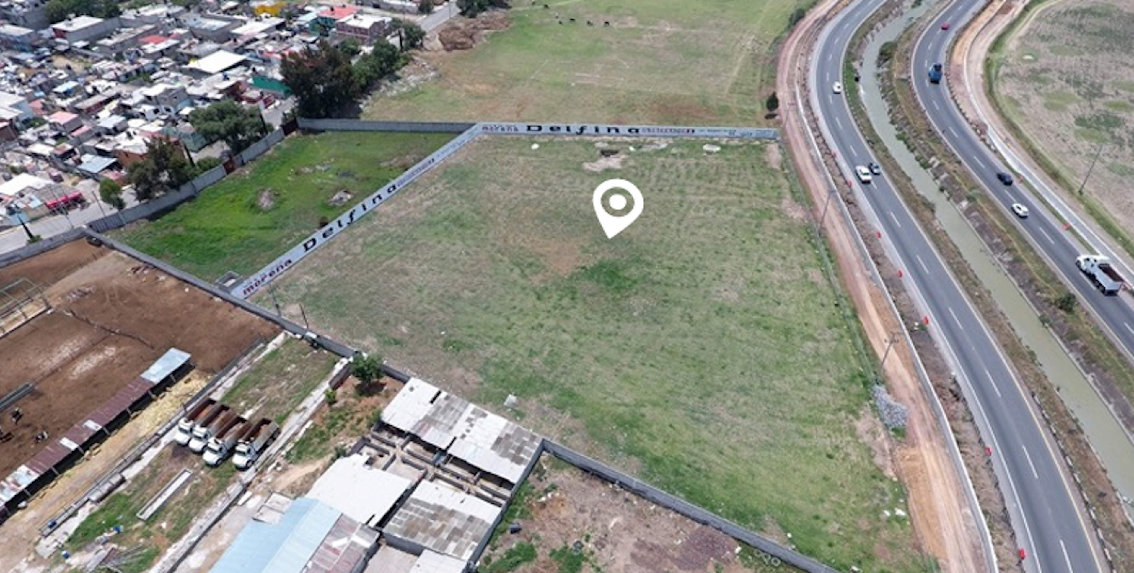 Vista aérea de un terreno vacío en una zona urbana, cercado y ubicado junto a una carretera. Ideal para desarrollo inmobiliario o comercial debido a su accesibilidad y visibilidad.