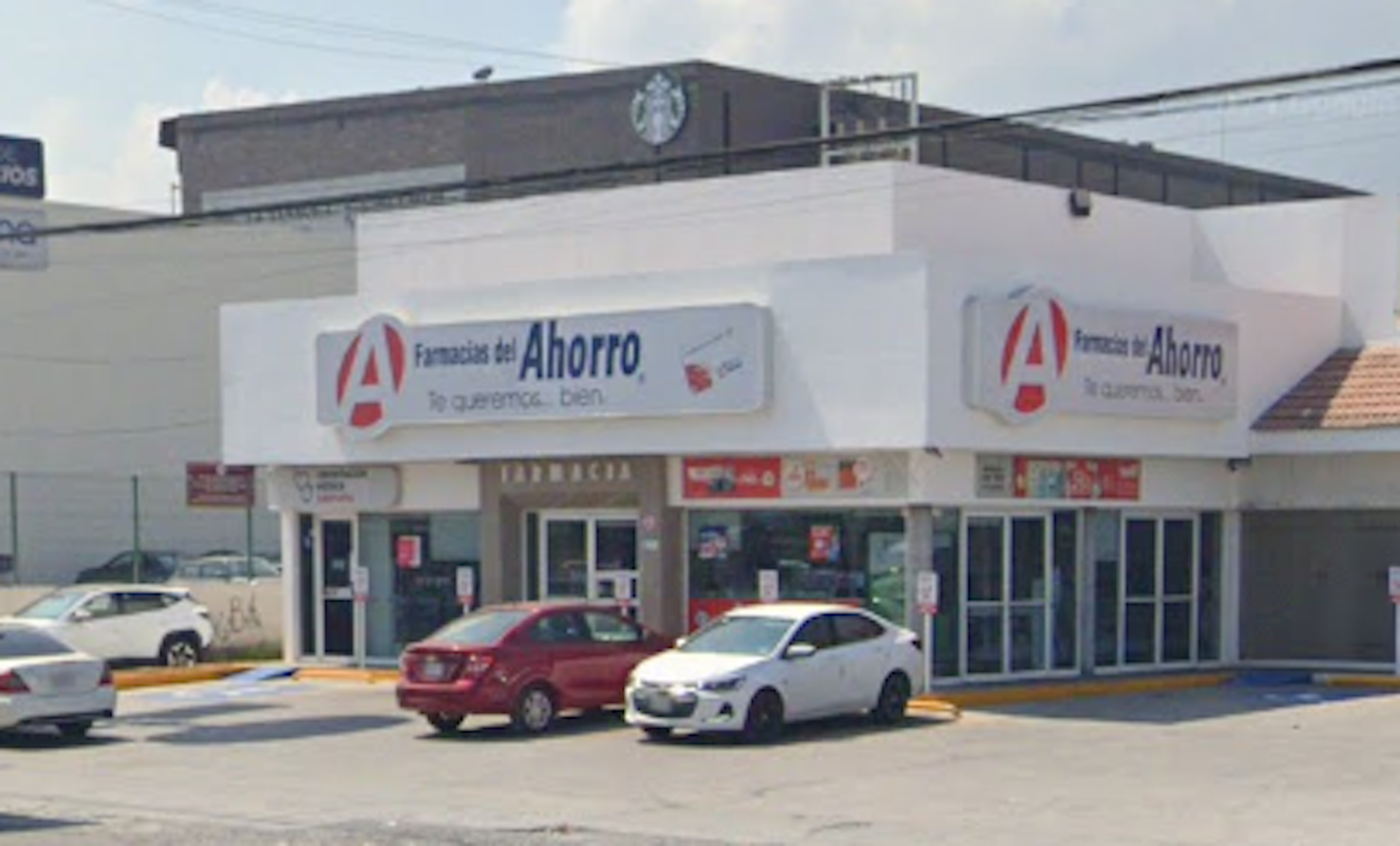 La imagen muestra una farmacia llamada Farmacias del Ahorro en un área comercial. Es un edificio blanco de un piso con grandes ventanales y letreros prominentes. Hay varios autos estacionados frente al local, indicando que es un negocio activo. Detrás se puede ver parte de un edificio más alto con el logo de Starbucks, sugiriendo que está en una zona comercial desarrollada.