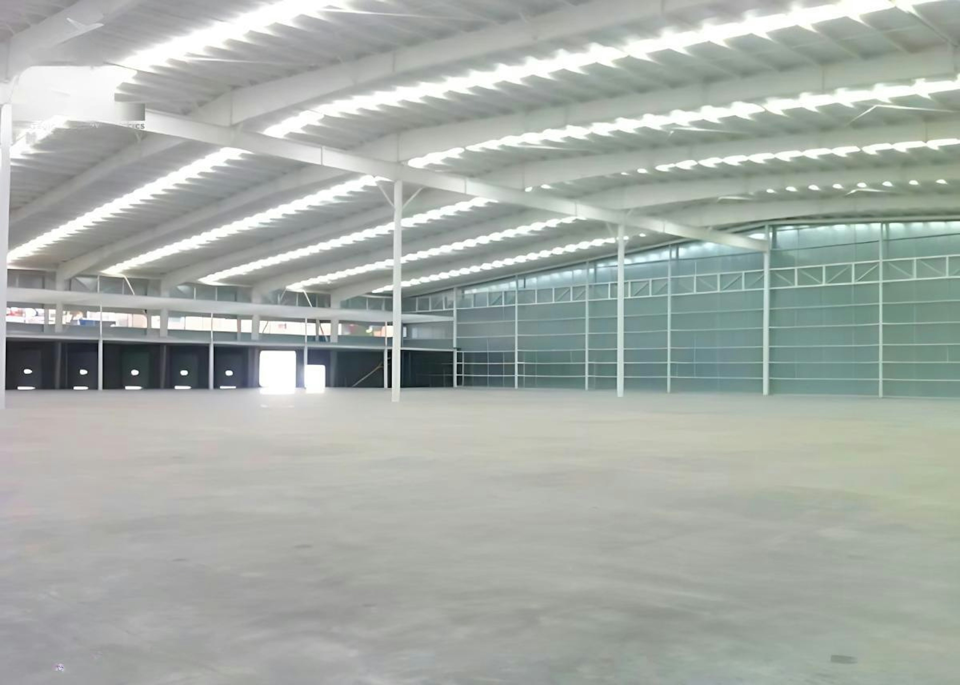 La imagen muestra un amplio espacio industrial interior con techo alto y estructura metálica. El área está bien iluminada con luces fluorescentes. Las paredes laterales son de vidrio, creando una sensación de apertura. El piso es de concreto liso, ideal para uso industrial o almacenamiento. Se observa un número de teléfono de contacto en la parte inferior, sugiriendo que el espacio está disponible para alquiler o venta.