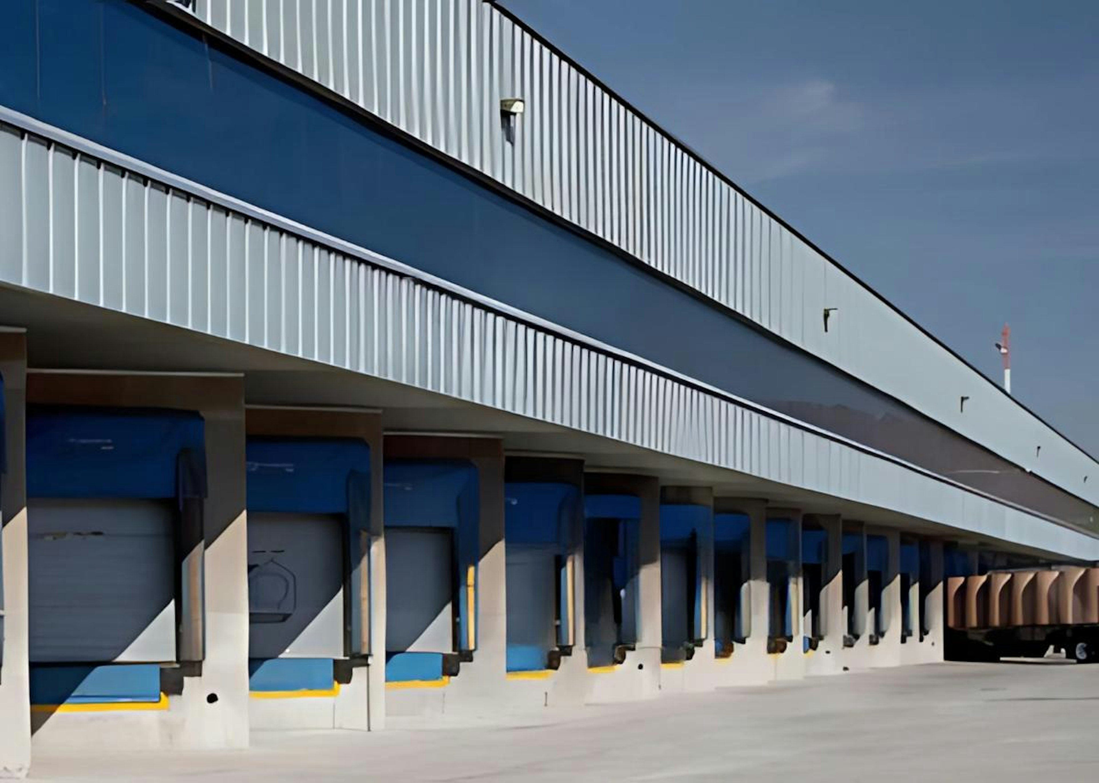 La imagen muestra una moderna instalación logística industrial con múltiples muelles de carga. La fachada combina colores azul y blanco, con un diseño arquitectónico contemporáneo. Se aprecia una serie de puertas de carga numeradas y una estructura elevada con barandillas. El letrero Strategic Industry Logistics sugiere que es un centro de distribución avanzado para operaciones industriales.