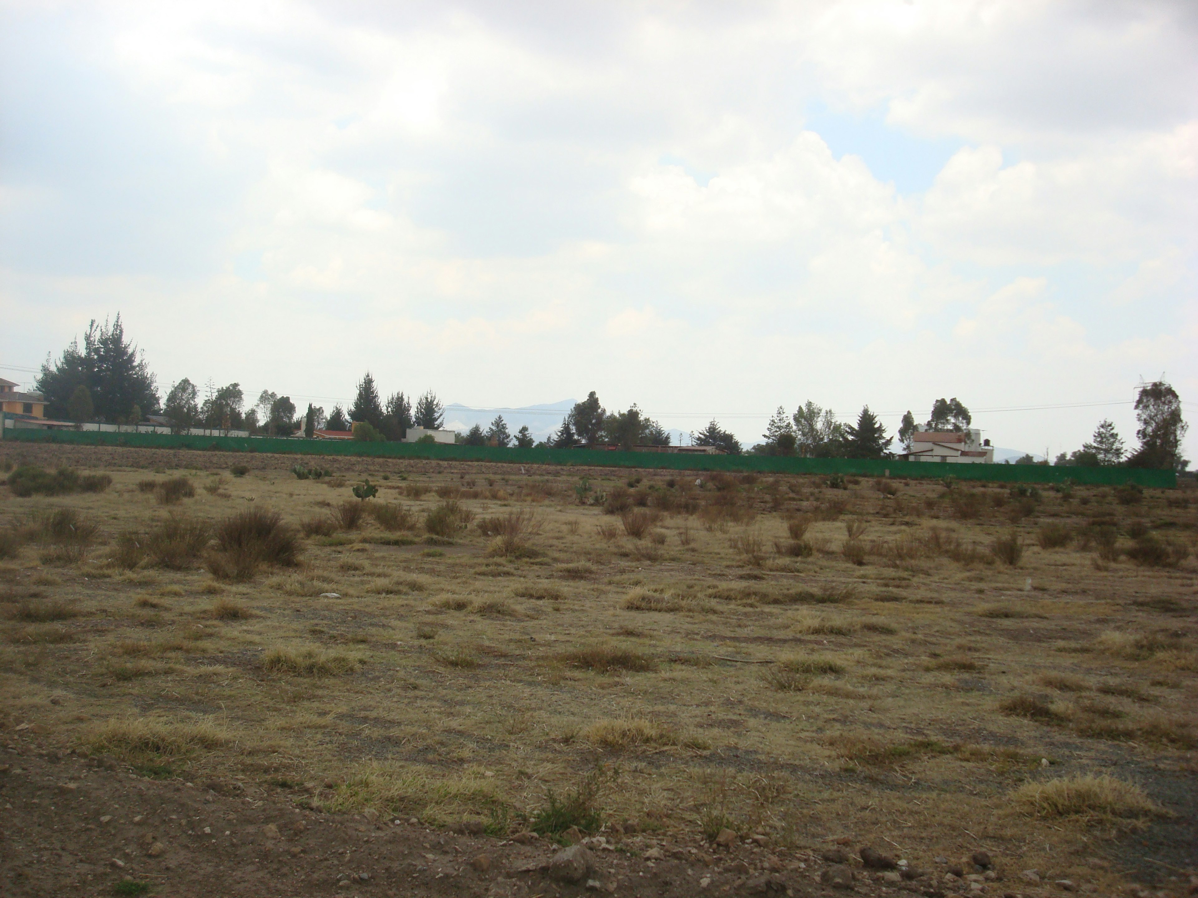 Terreno amplio y despejado, potencial para desarrollo inmobiliario o agrícola. Cielo parcialmente nublado y vegetación baja. Zona tranquila con edificaciones a lo lejos.