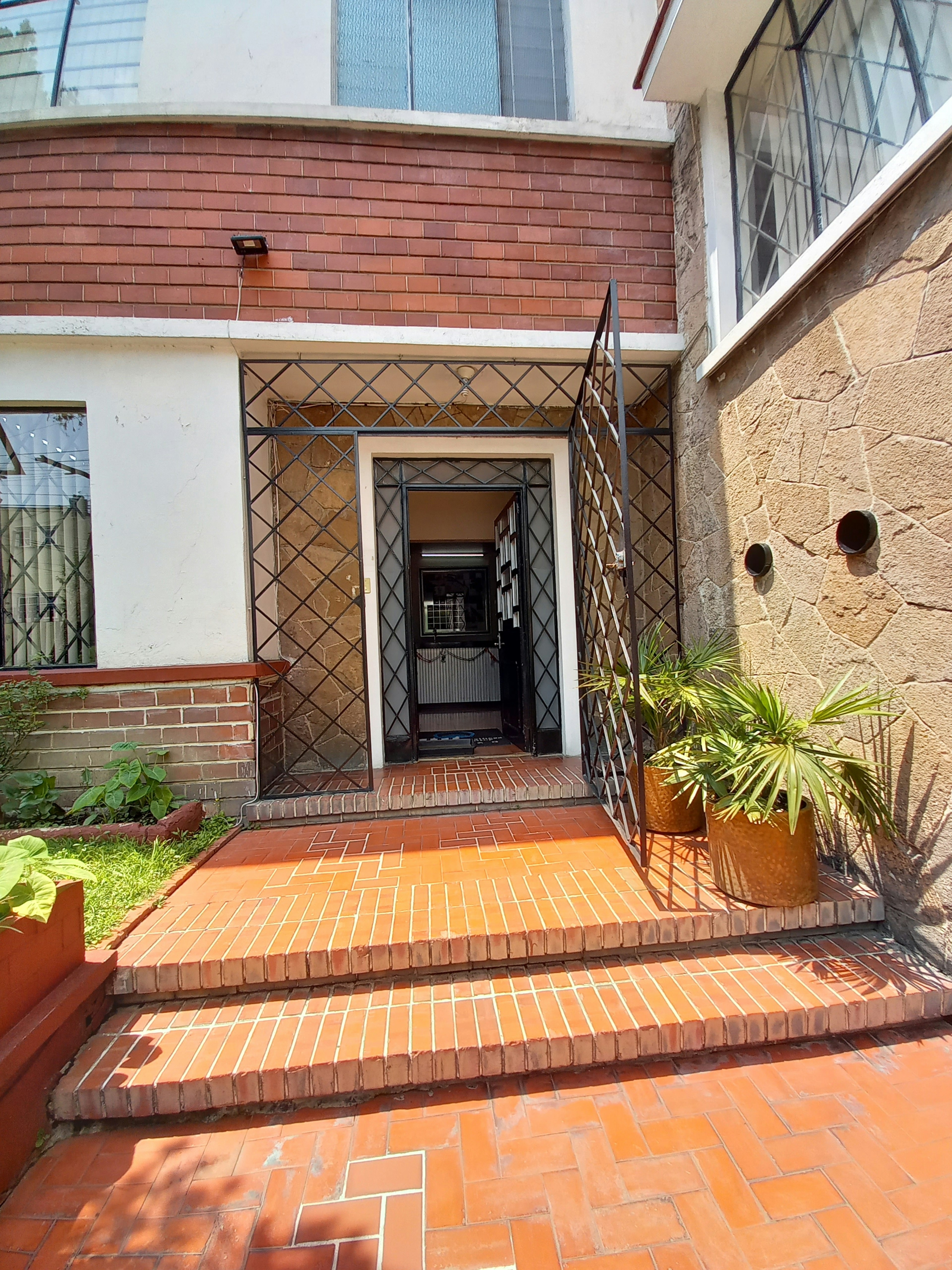 Entrada de una casa con escalones de ladrillo y puerta de metal. Fachada con combinación de ladrillo y piedra, ventanas con vidrios esmerilados y plantas decorativas.