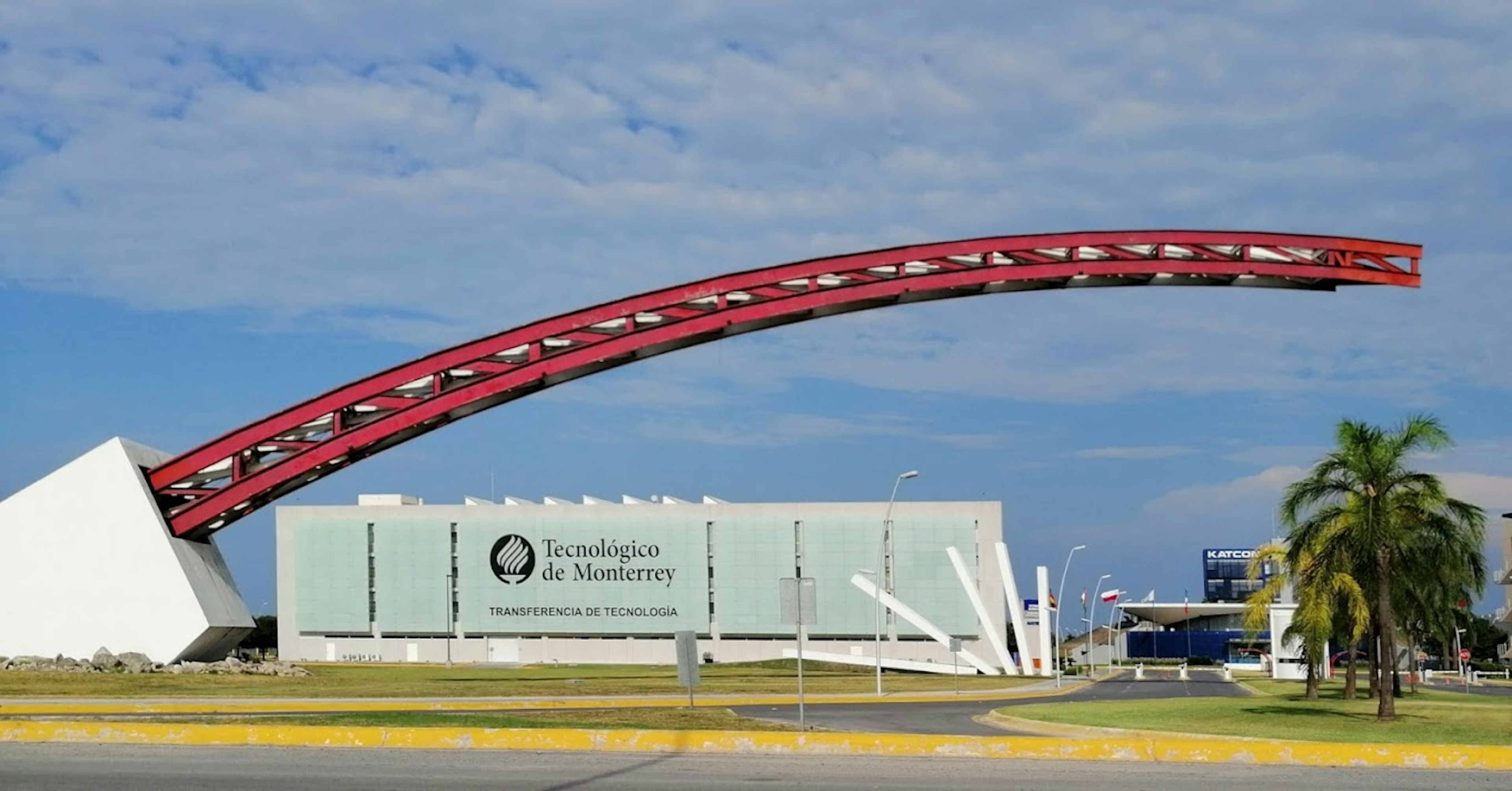 La imagen muestra la entrada del campus del Tecnológico de Monterrey, una prestigiosa institución educativa en México. Se aprecia un edificio moderno de color blanco y azul claro con el nombre Tecnológico de Monterrey visible. Destaca un gran arco rojo que forma parte de la estructura arquitectónica, creando un diseño llamativo y contemporáneo. El entorno incluye palmeras y un cielo azul con nubes, dando una sensación de ambiente cálido y acogedor.