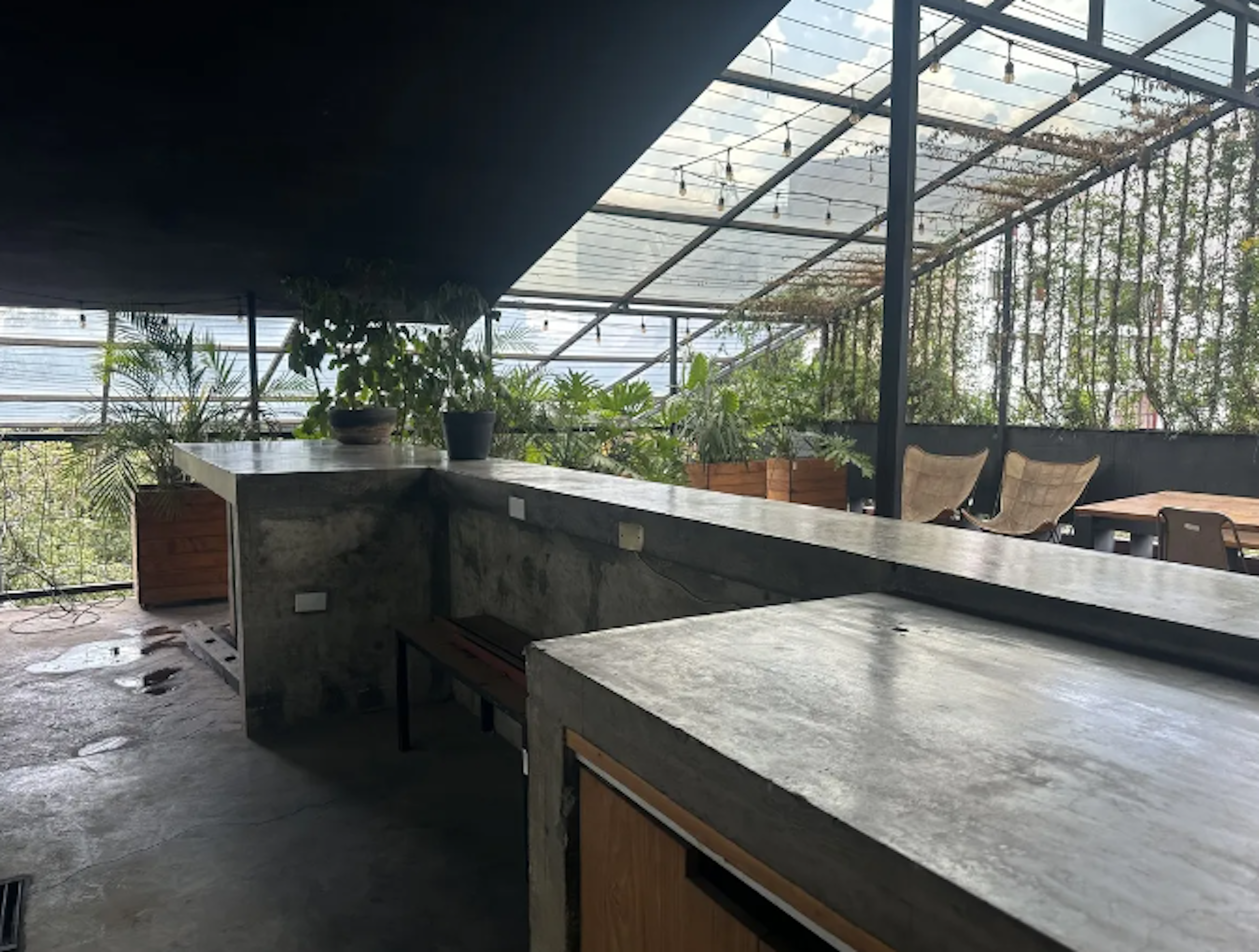 Esta imagen muestra un espacio interior moderno con techo acristalado, ideal para un restaurante o área común de un complejo residencial de lujo. Cuenta con una barra de concreto, vegetación abundante, y una vista al exterior a través de paneles de vidrio. El diseño combina elementos industriales con toques naturales, creando un ambiente acogedor y sofisticado.