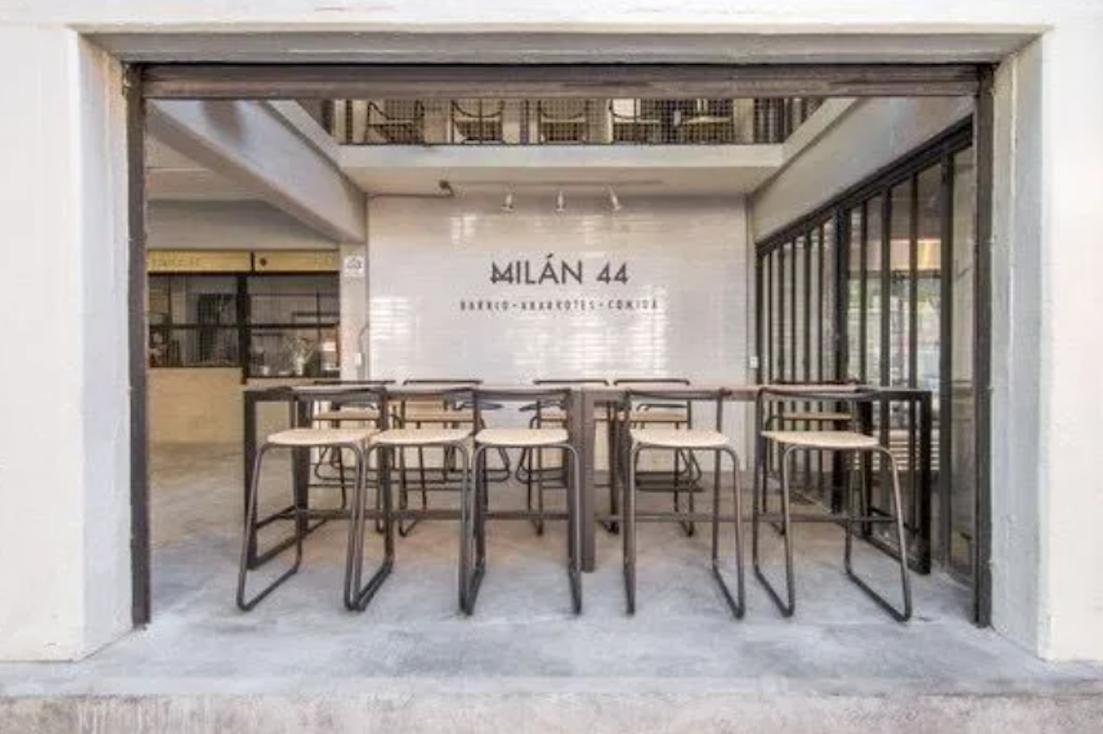 La imagen muestra la entrada de un establecimiento llamado MILÁN 44. Es un espacio moderno y minimalista con un diseño industrial. La entrada tiene mesas altas y taburetes, ideal para un café o bar. El interior parece espacioso, con un techo alto y una mezzanine visible. El estilo es limpio, con colores neutros y materiales como metal y concreto, típico de espacios comerciales renovados en zonas urbanas.