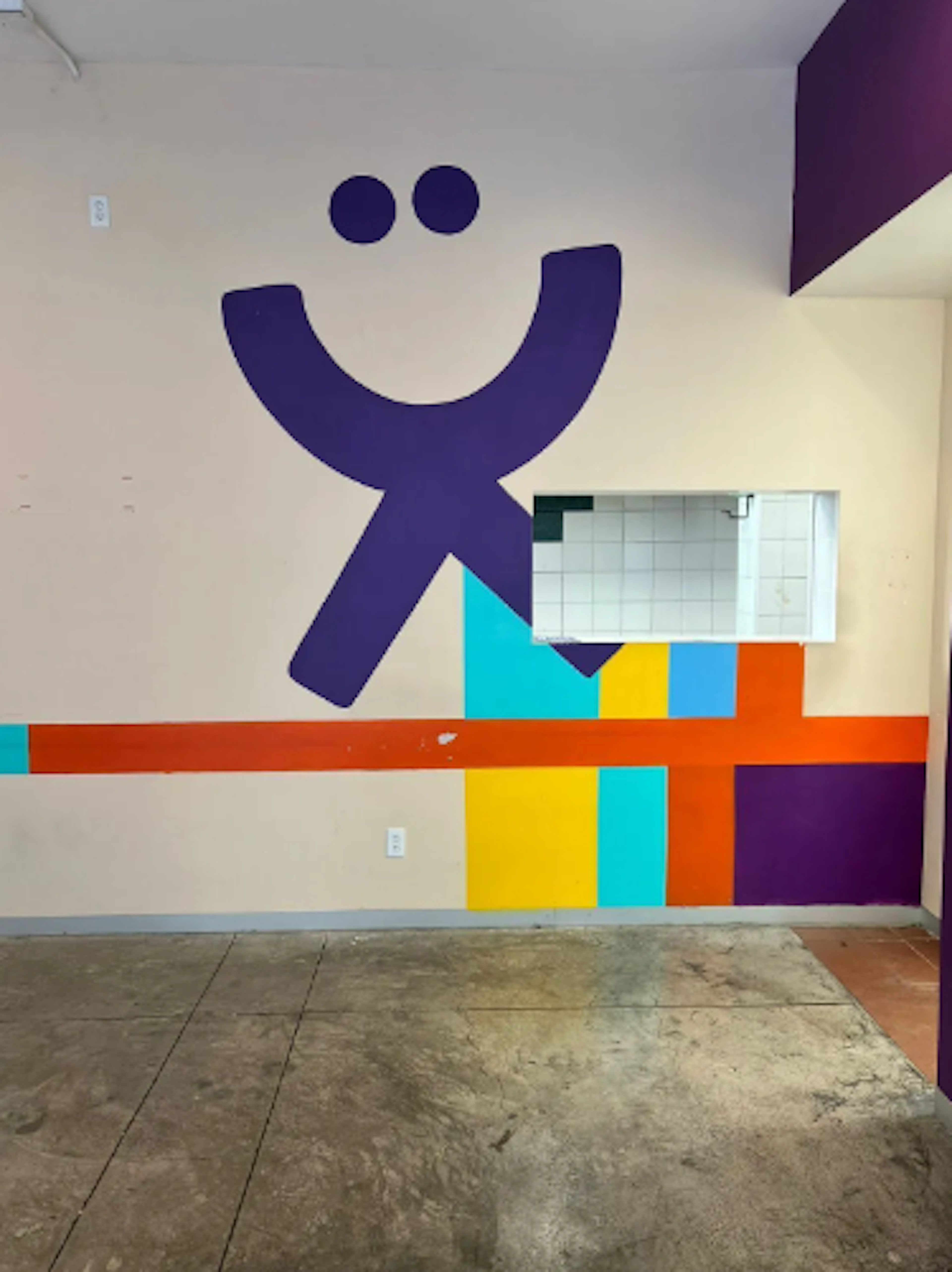 La imagen muestra un espacio interior con una pared decorada con un gran logo morado en forma de figura sonriente y bloques de colores brillantes. Hay un espejo rectangular integrado en el diseño. El suelo es de baldosas y la pared principal es de color crema. Este diseño colorido y alegre podría pertenecer a una guardería, centro infantil o área recreativa dentro de un edificio residencial o comercial.