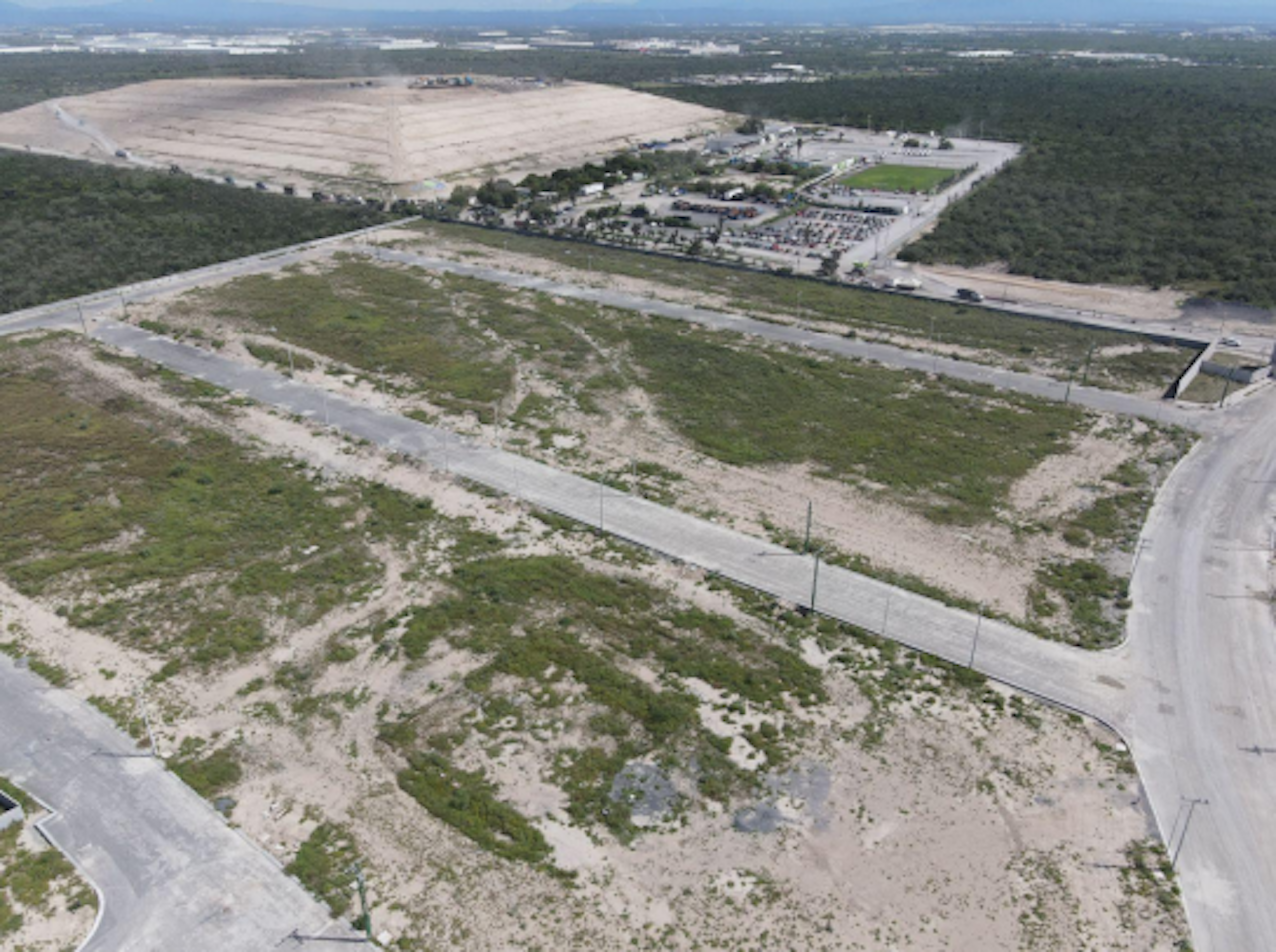 La imagen muestra una vista aérea de un área en desarrollo, con grandes extensiones de terreno despejado y algunas áreas pavimentadas. Se observa una zona industrial o comercial en el fondo con edificios y estacionamientos. El paisaje es principalmente árido, con vegetación dispersa. Hay carreteras que dividen las parcelas, sugiriendo una planificación para futura urbanización o expansión industrial.