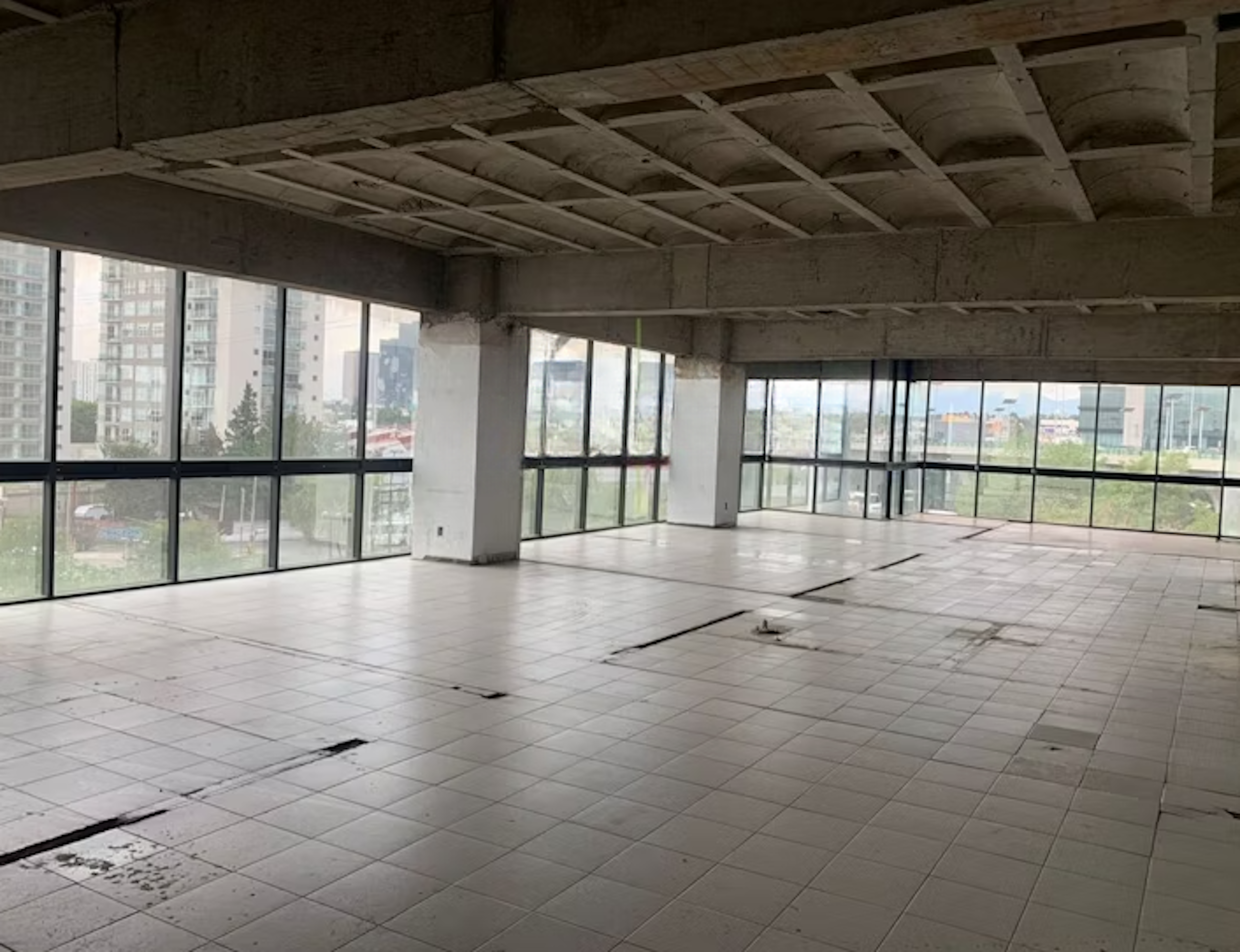 La imagen muestra un amplio espacio comercial en construcción o renovación. Tiene grandes ventanales que ofrecen vistas panorámicas de la ciudad circundante, con edificios altos visibles en el exterior. El interior está inacabado, con un techo expuesto y un piso de baldosas. El espacio parece ideal para una oficina de planta abierta o un local comercial de gran tamaño, con mucha luz natural y potencial para personalización.