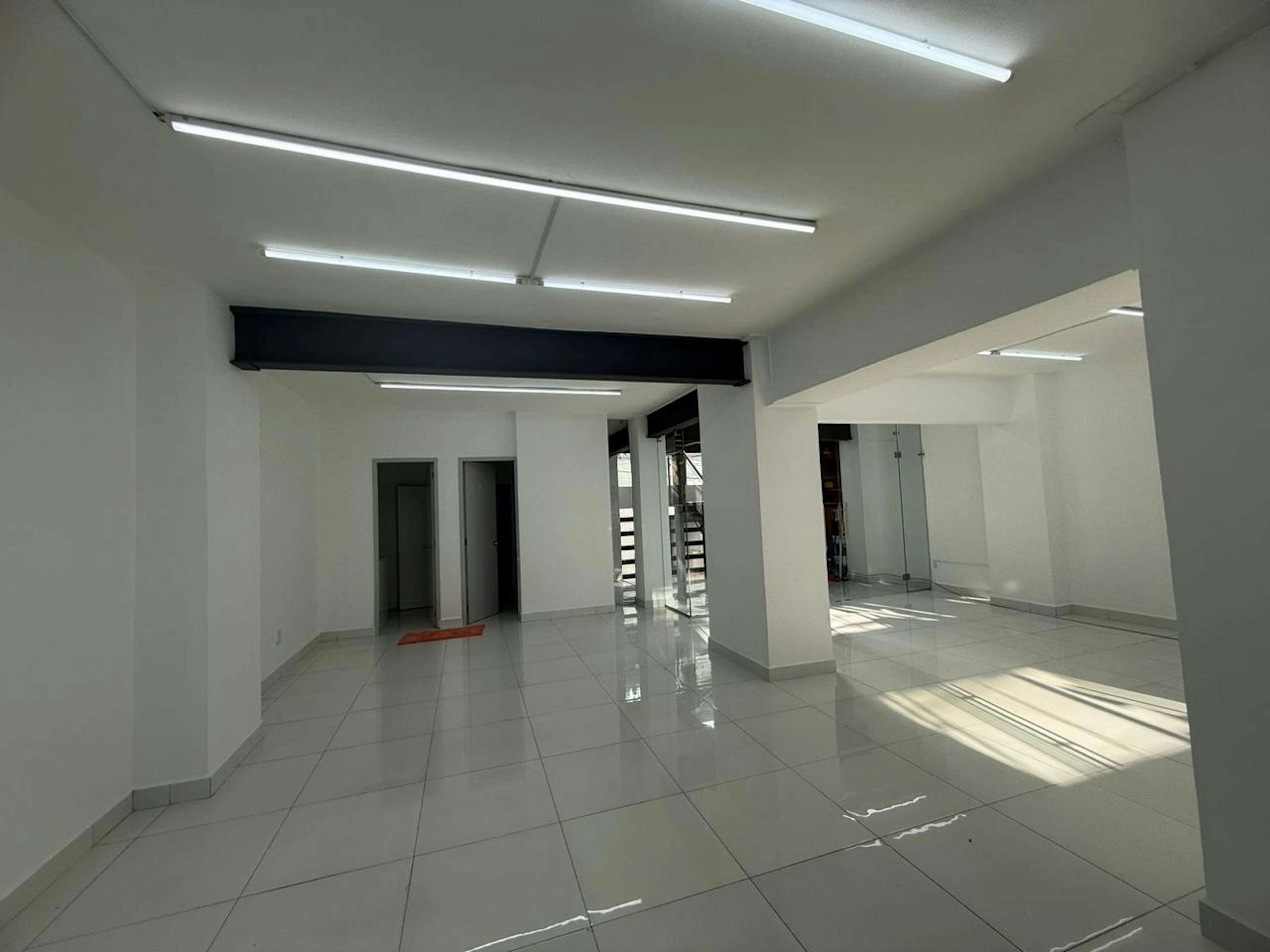 La imagen muestra un espacio interior amplio y luminoso, ideal para oficinas o locales comerciales. Cuenta con paredes blancas, suelo de baldosas brillantes y luz natural que entra por ventanas. Se observan varias puertas, una zona abierta y un área con divisiones de vidrio. La iluminación artificial proviene de tubos fluorescentes en el techo. El diseño es moderno y minimalista, ofreciendo versatilidad para diversos usos comerciales.