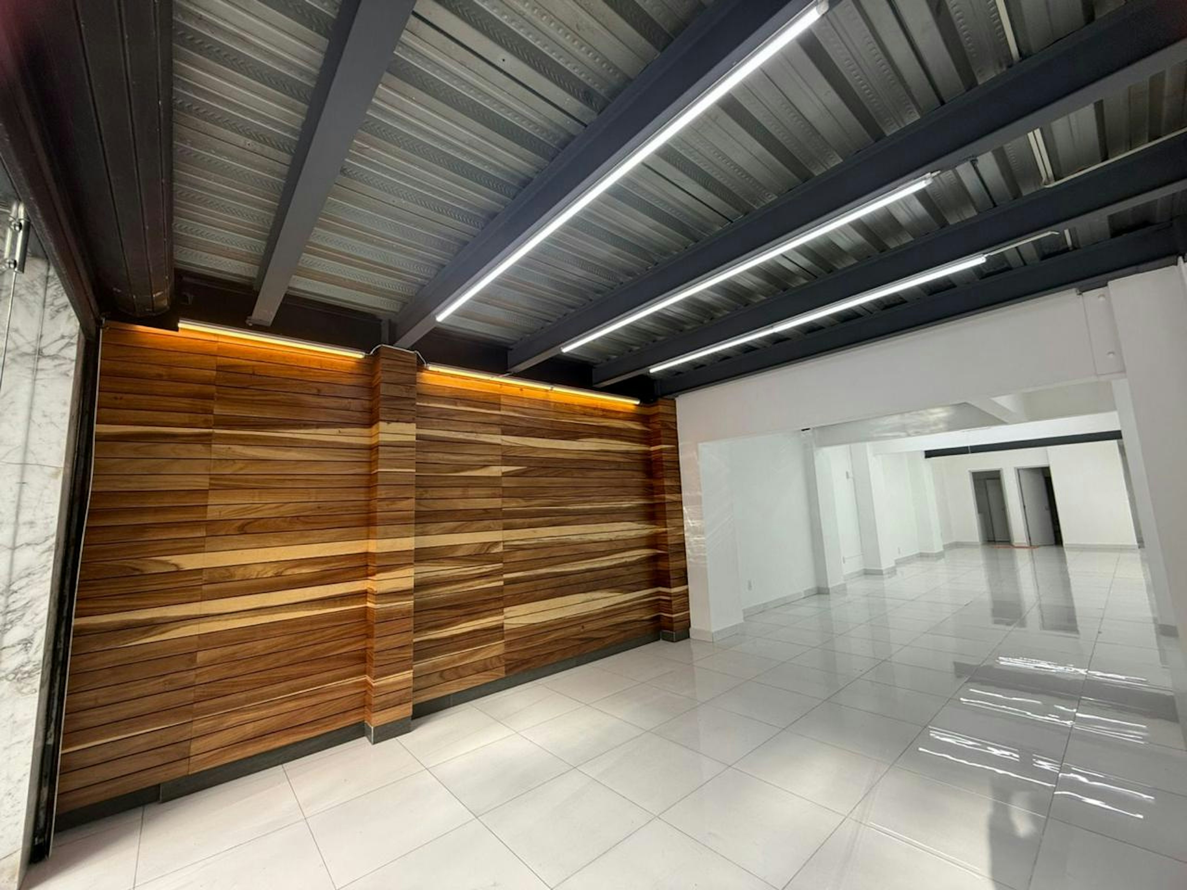 La imagen muestra un espacio interior moderno y elegante. Destaca una pared de madera con iluminación cálida en la parte superior. El techo tiene vigas expuestas con luces LED lineales. El piso es de baldosas blancas brillantes. A la derecha se ve un pasillo blanco que conduce a otras áreas. El diseño combina elementos industriales con acabados cálidos, creando un ambiente contemporáneo y acogedor, ideal para un lobby o área de recepción en un edificio comercial o residencial de lujo.