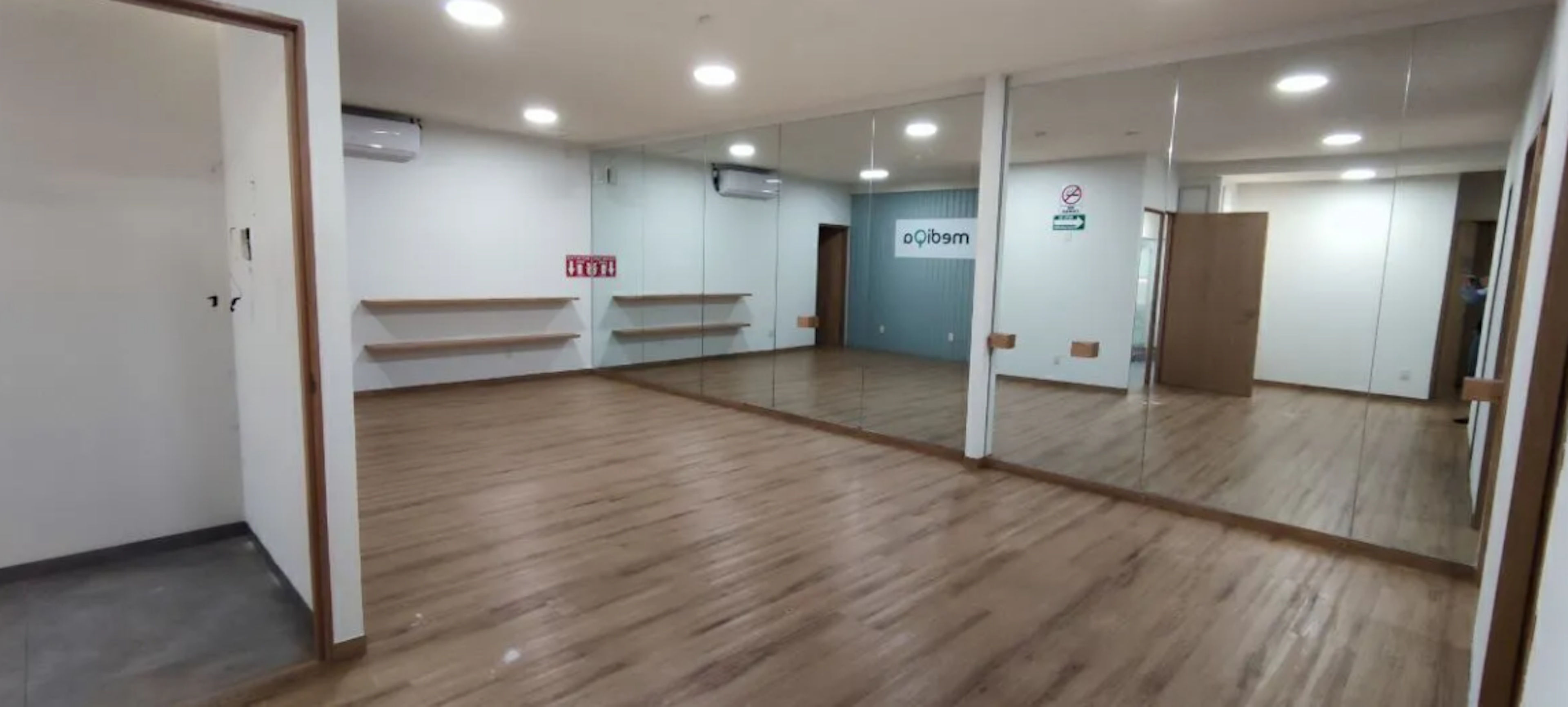 La imagen muestra un espacio interior amplio y luminoso, probablemente un estudio de danza o fitness. Tiene un piso de madera pulida, grandes espejos en una pared, iluminación LED empotrada en el techo, y aire acondicionado. Hay estantes montados en la pared y una puerta visible. El espacio parece estar recién renovado, con paredes blancas limpias y un aspecto moderno general, ideal para actividades físicas o clases.