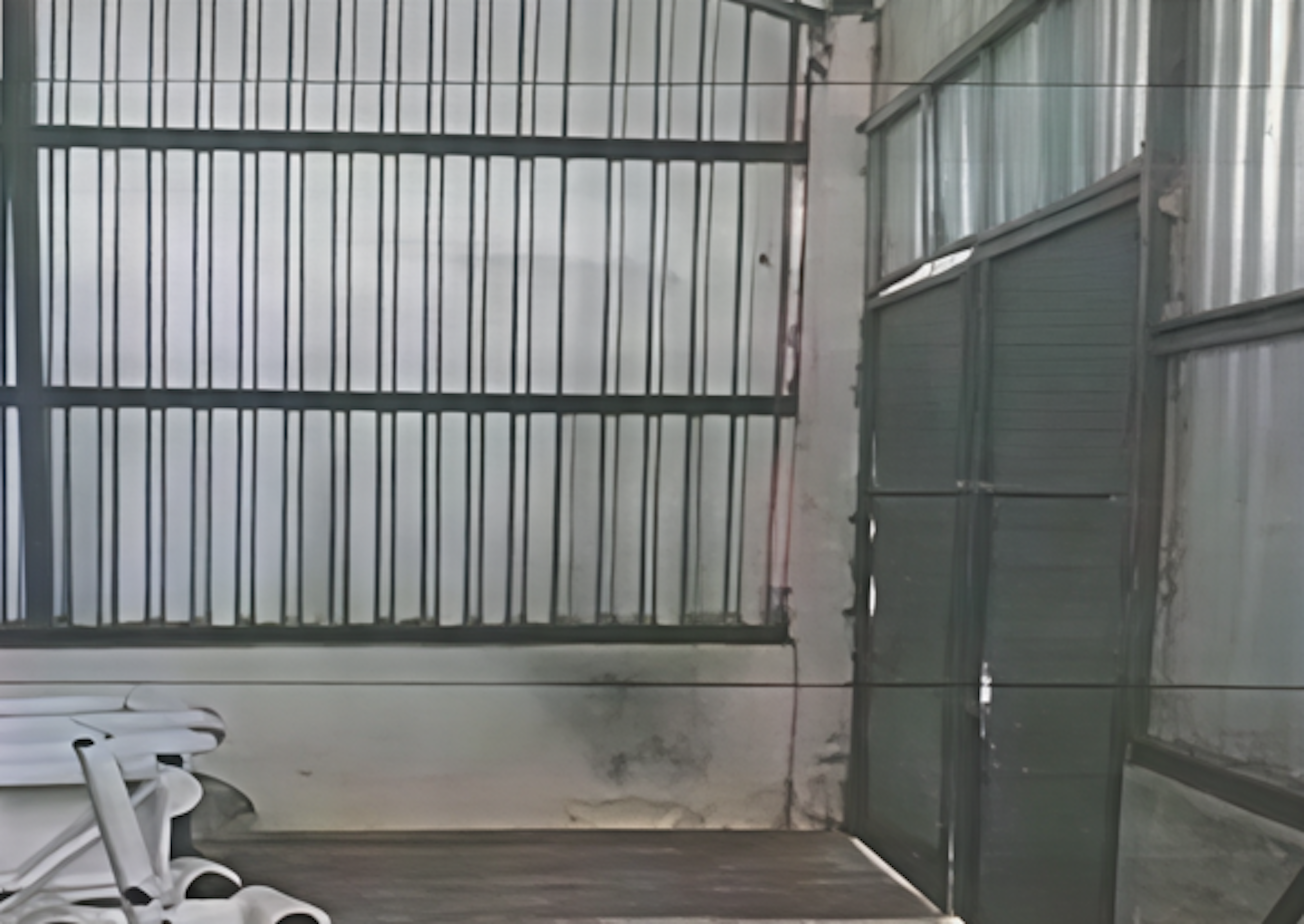 La imagen muestra un espacio interior con aspecto industrial o de almacén. Se observa una pared con barrotes metálicos que podría ser una puerta o división. A la derecha hay una estructura verde que parece ser otra puerta o ventana. El suelo es de concreto y las paredes están en mal estado, con manchas y desgaste visible. Este espacio podría ser parte de un edificio industrial o un almacén que necesita renovación.