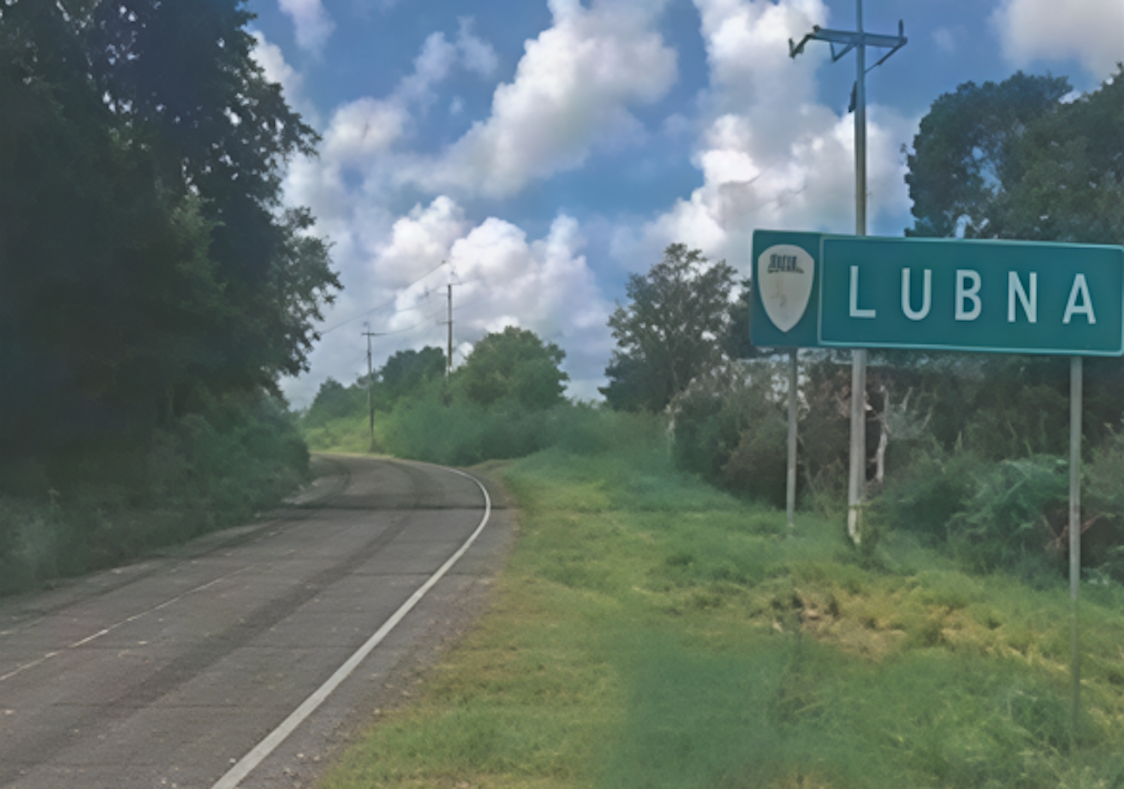 La imagen muestra una carretera rural que conduce a la ciudad de Lubna, como lo indica un letrero verde al borde del camino. El paisaje es exuberante y verde, con árboles y vegetación a ambos lados de la carretera. El cielo es azul con nubes blancas esponjosas. La escena sugiere una zona residencial tranquila o potencialmente en desarrollo, ideal para propiedades rurales o suburbanas.