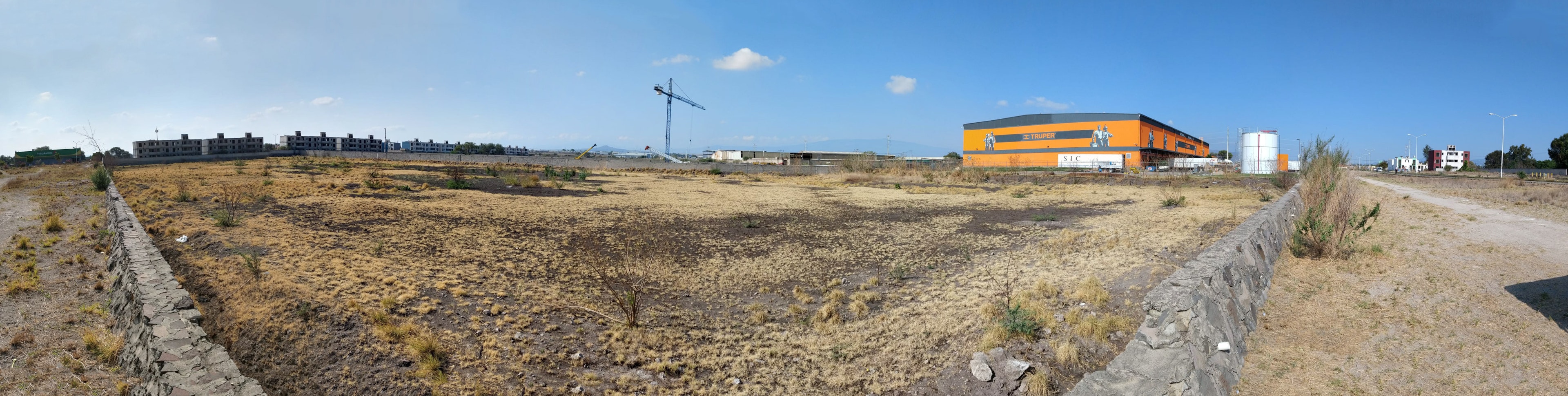 La imagen muestra un terreno baldío en una zona en desarrollo. Se observa un área extensa de tierra árida y seca, con algunos arbustos dispersos. En el fondo se pueden ver edificios en construcción, una grúa, y una estructura naranja que parece ser un almacén o instalación industrial. Hay un muro de piedra que delimita parte del terreno. El paisaje sugiere una zona suburbana o industrial en expansión, potencialmente atractiva para futuros desarrollos inmobiliarios.