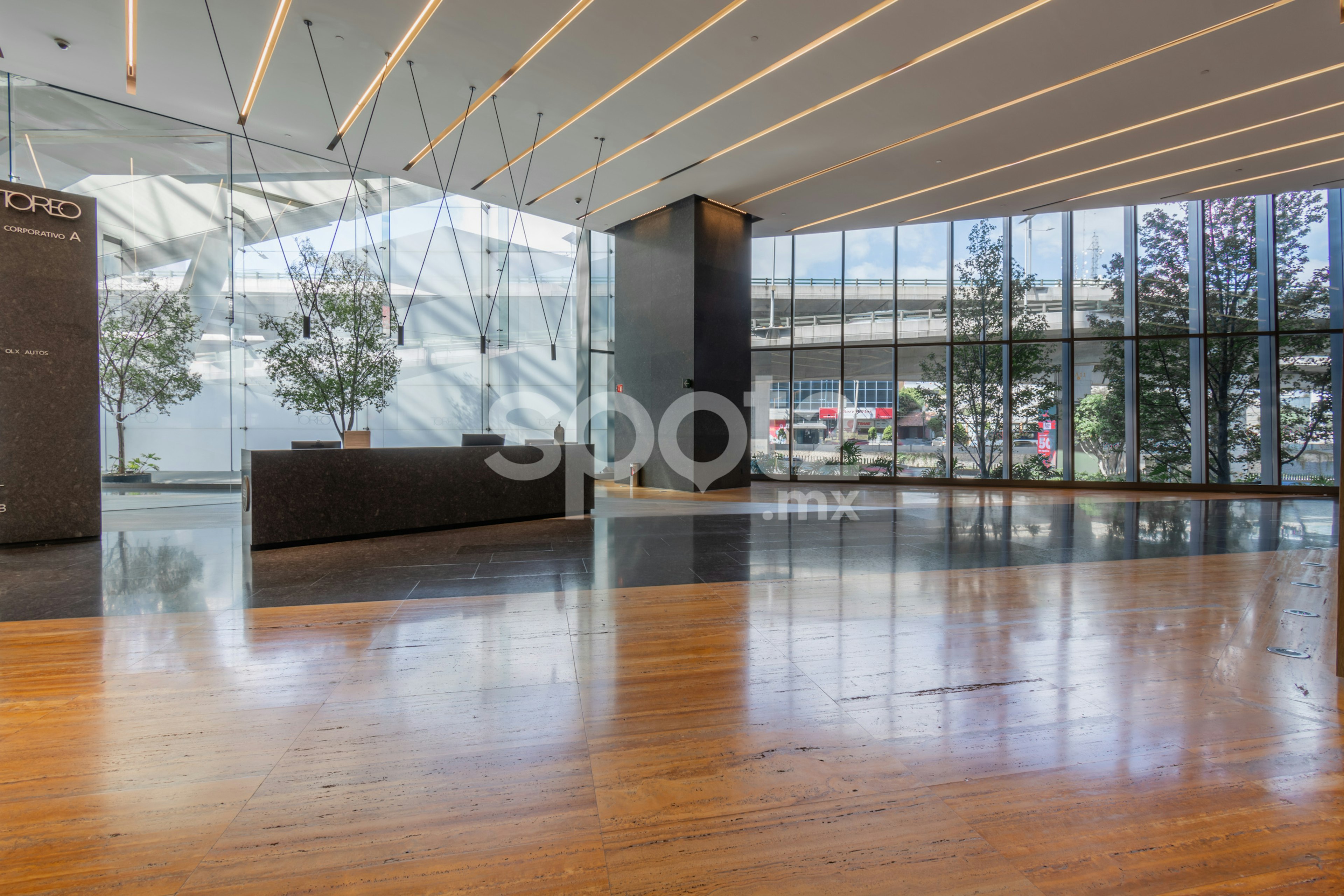 La imagen muestra un elegante vestíbulo de un edificio corporativo moderno. Cuenta con un techo con iluminación lineal, un piso de madera pulida, grandes ventanales que permiten la entrada de luz natural y vistas al exterior. Se aprecia una recepción de diseño minimalista y plantas decorativas. El espacio transmite una sensación de amplitud, luminosidad y sofisticación, ideal para una oficina de alto nivel.