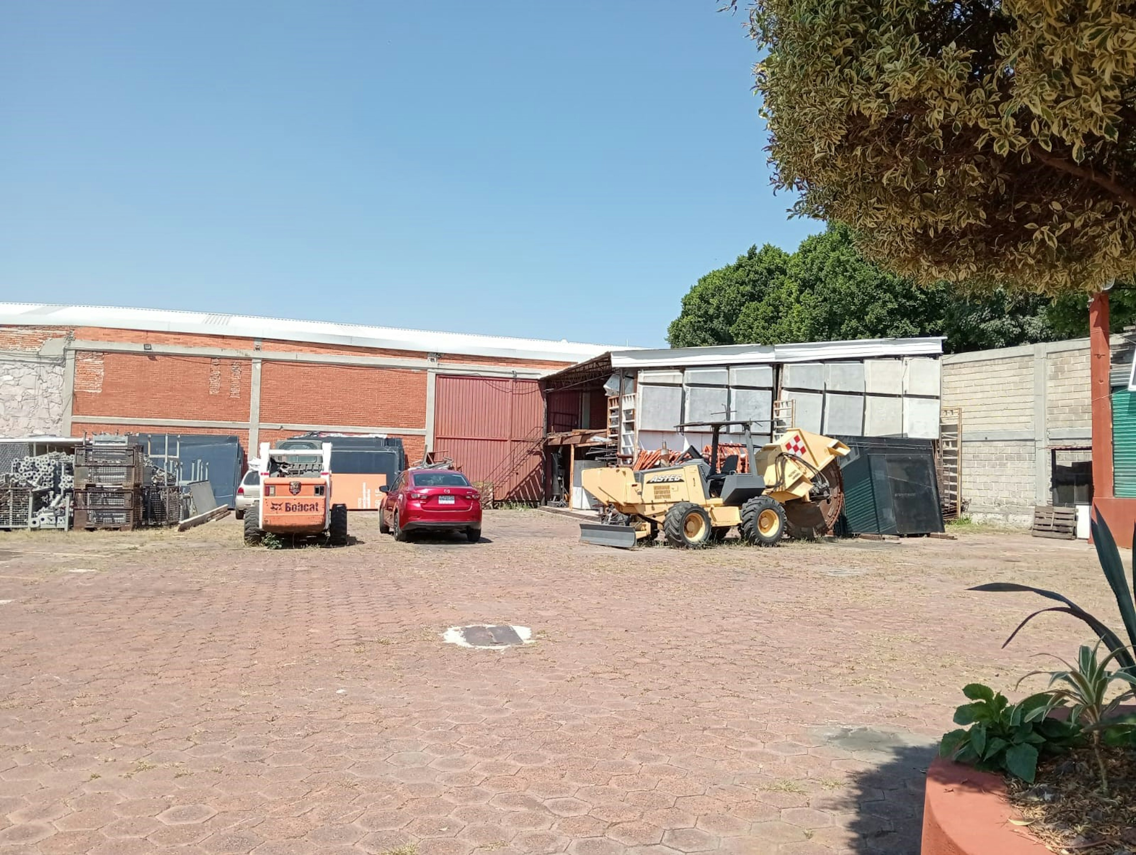 La imagen muestra un patio industrial o área de almacenamiento exterior. Se observan edificios de ladrillo y estructuras metálicas, junto con vehículos y maquinaria pesada como un Bobcat y un tractor. El suelo es de adoquines y hay diversos materiales y equipos almacenados. Este espacio podría ser parte de un complejo industrial o un área de almacenamiento para una empresa de construcción o logística.