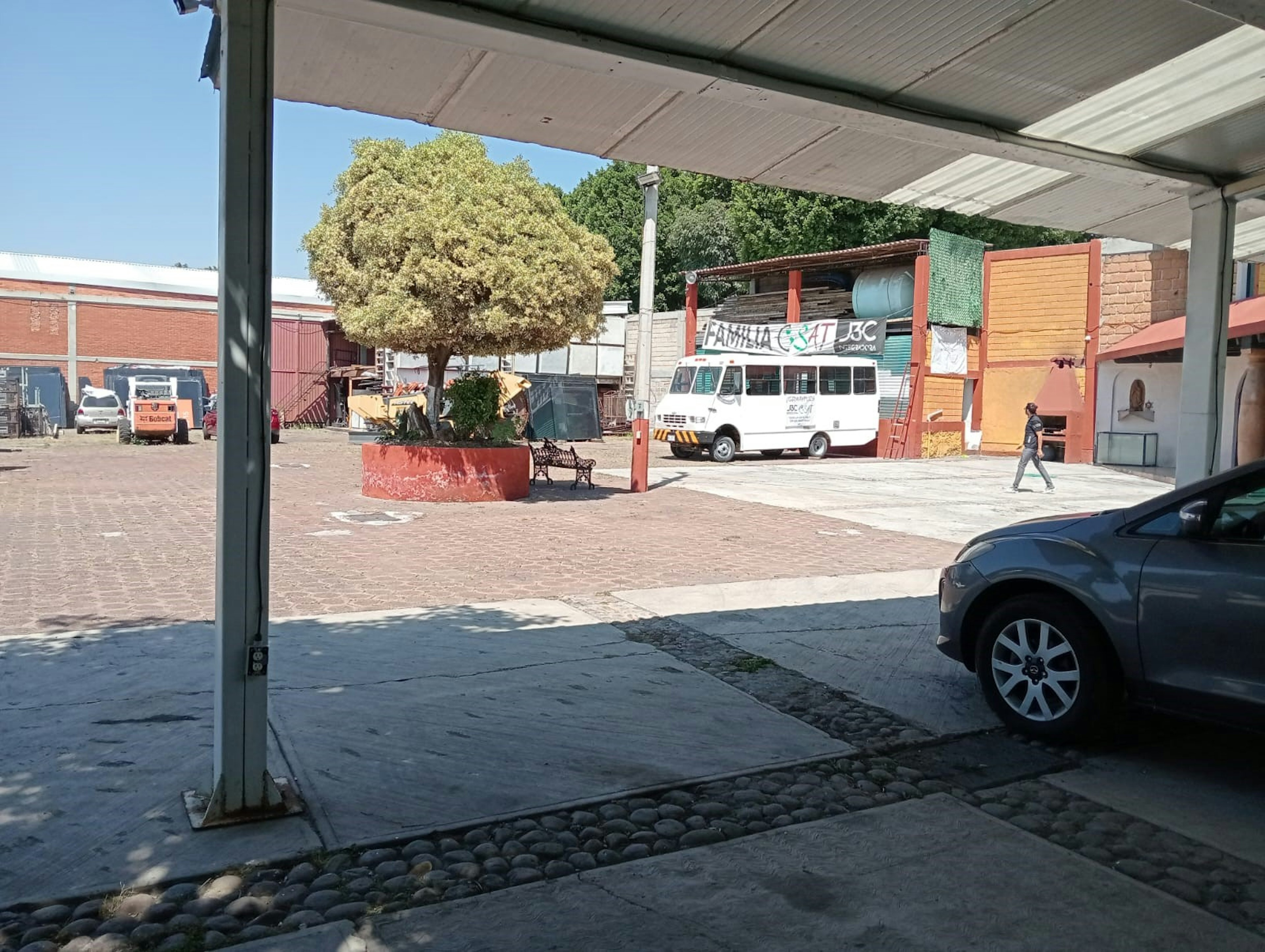 La imagen muestra un área semi-industrial con un patio pavimentado. Se ve un gran árbol en una jardinera roja, un minibús blanco con un letrero, y varios edificios de ladrillo. Hay un techo visible en primer plano, sugiriendo que la foto se tomó desde algún tipo de estructura cubierta. El espacio parece ser una mezcla de usos comerciales y posiblemente residenciales, con potencial para desarrollo o remodelación.