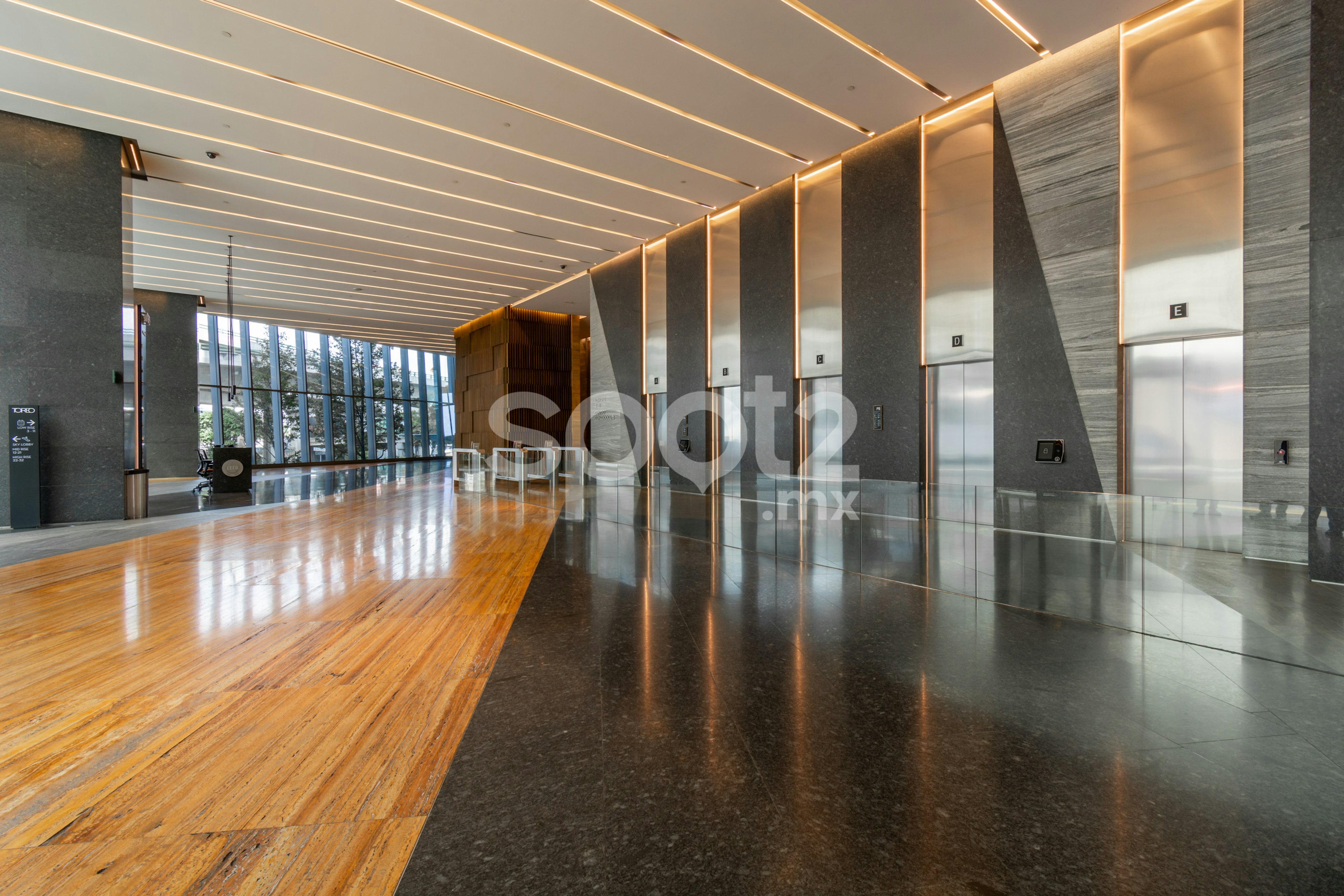 This image showcases a modern, high-end office lobby or commercial building interior. It features a sleek design with linear ceiling lights, polished floors combining wood and stone, and a row of elevators. The space exudes luxury and professionalism with its clean lines, ambient lighting, and expansive windows offering natural light. The Scout watermark suggests this is a professional real estate or architectural photograph.