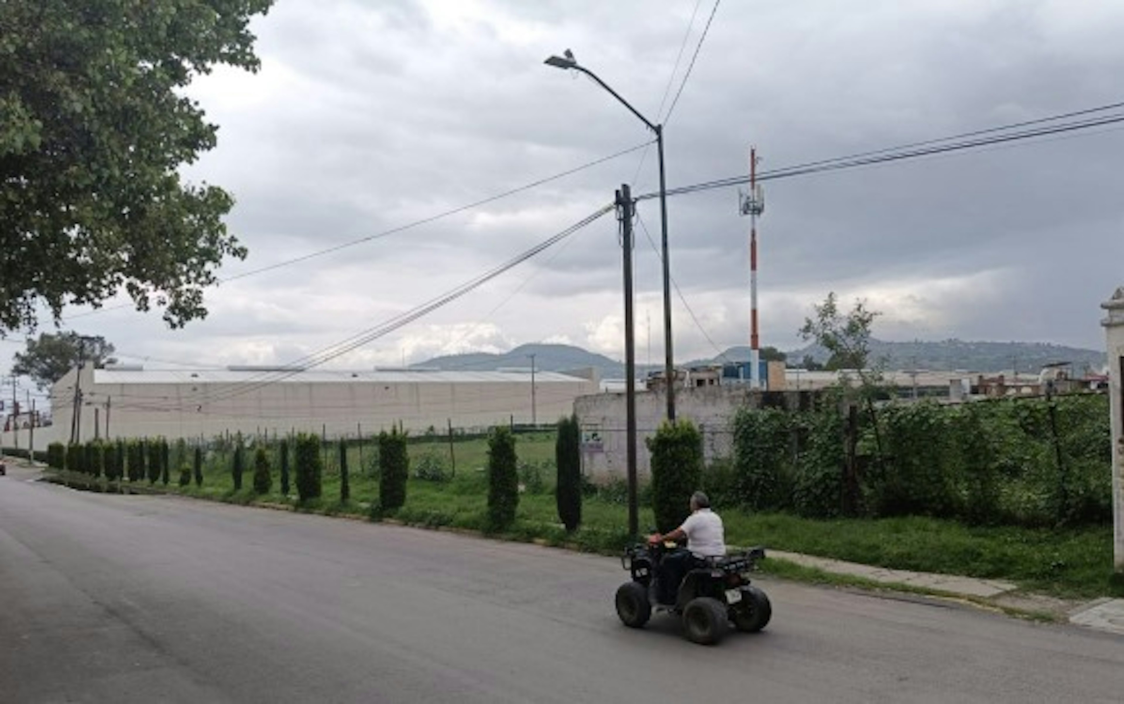 La imagen muestra una zona suburbana con una calle pavimentada bordeada por una hilera de árboles y arbustos. Al fondo se observa una gran estructura blanca, posiblemente un almacén o edificio industrial. El paisaje incluye montañas en la distancia y un cielo nublado. La presencia de infraestructura eléctrica y un individuo en un vehículo todo terreno sugiere una área en desarrollo o transición.