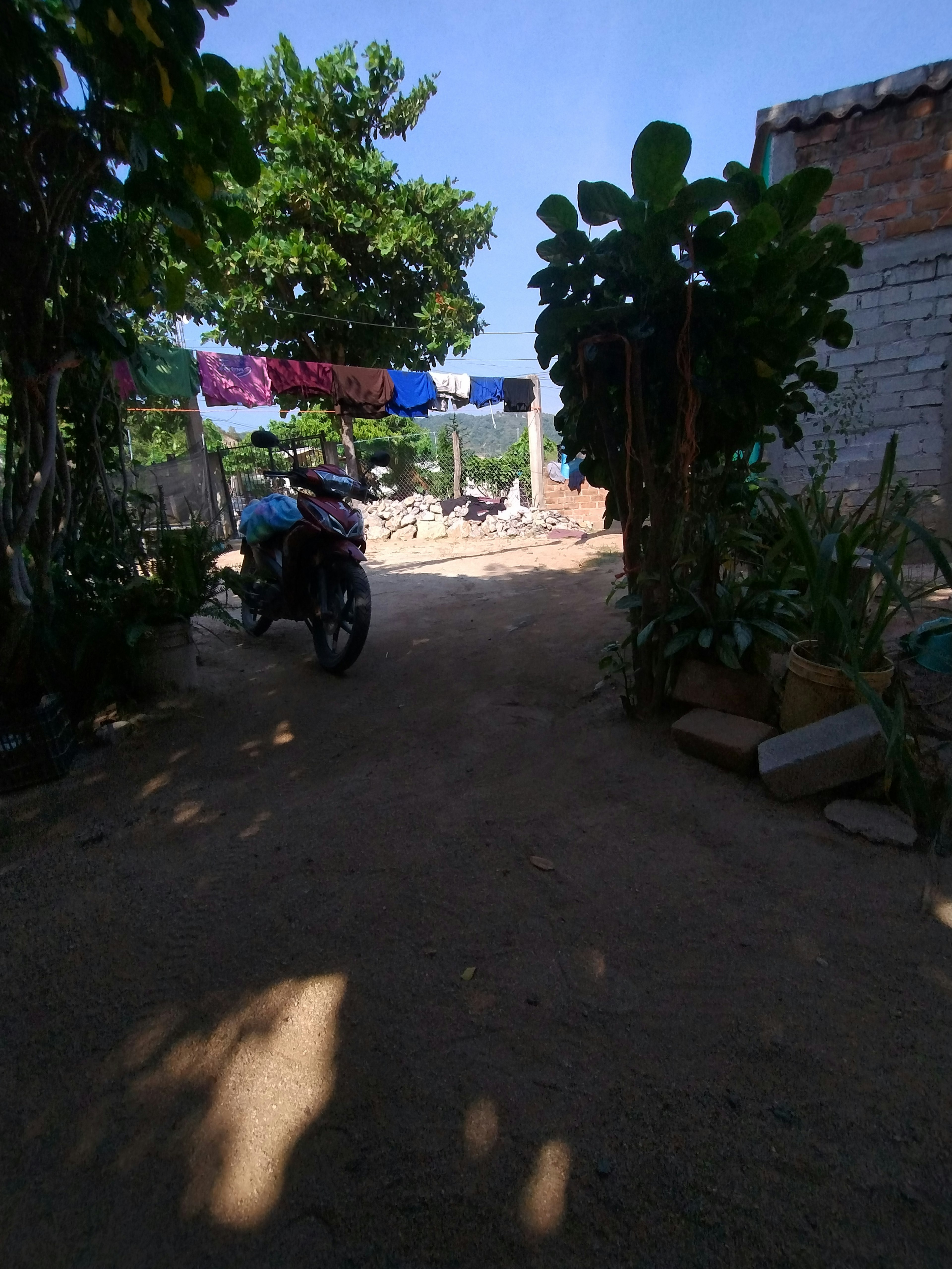 La imagen muestra un patio trasero rústico con suelo de tierra, rodeado de vegetación y árboles. Hay ropa tendida en una cuerda, una motocicleta estacionada y una pared de ladrillo parcialmente construida. El ambiente sugiere una propiedad en una zona rural o suburbana en desarrollo, con un estilo de vida sencillo y práctico. El espacio exterior parece ser utilizado para múltiples propósitos, incluyendo lavandería y estacionamiento.