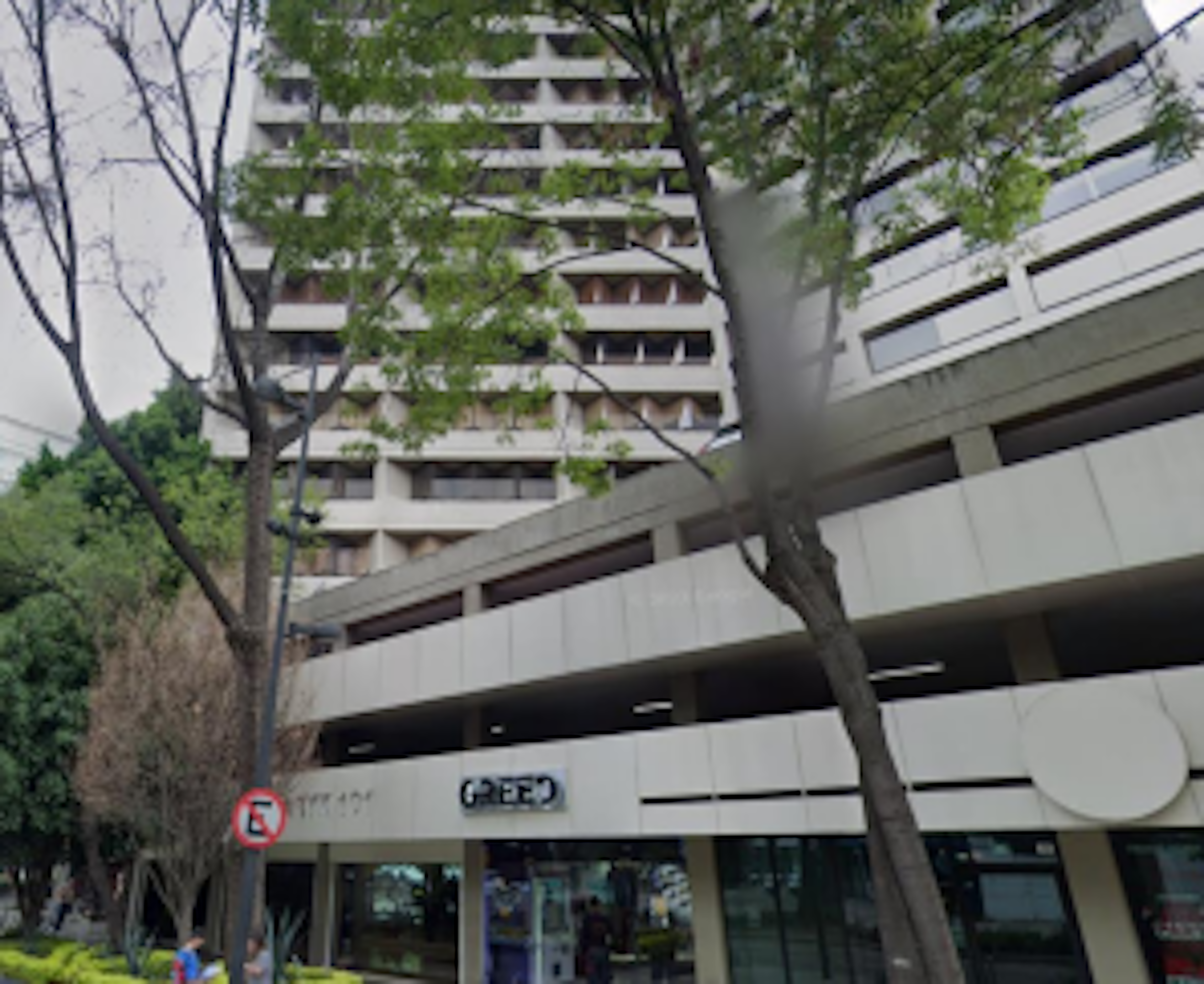 La imagen muestra un edificio residencial de varios pisos con balcones. La fachada es de color claro y tiene una estructura moderna. En la planta baja hay locales comerciales, incluyendo uno con el letrero OXXO. El edificio está rodeado de árboles que proporcionan sombra y verdor al entorno urbano. Se aprecia una señal de tráfico que prohíbe el estacionamiento. La arquitectura sugiere que es un complejo de apartamentos en una zona urbana.