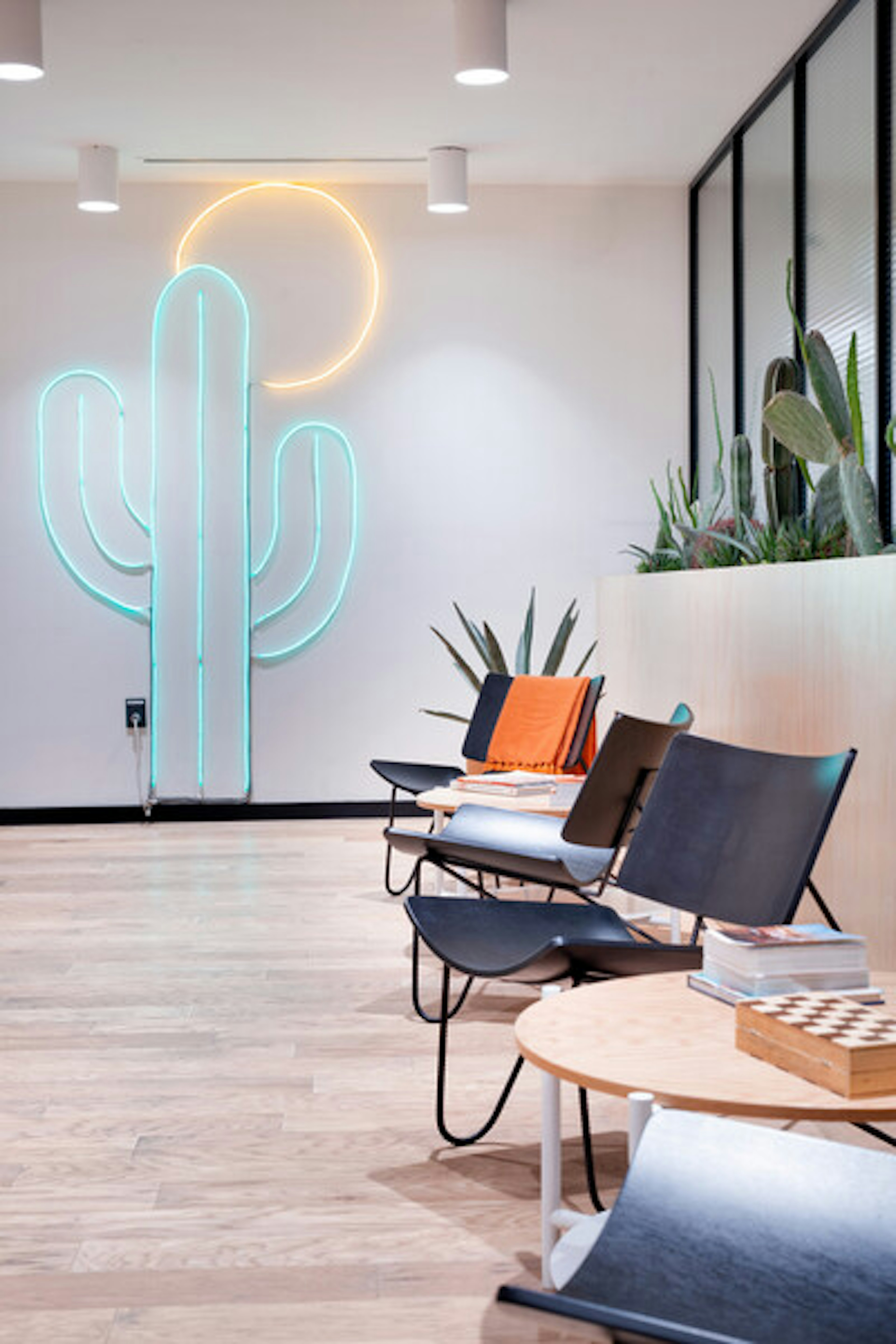 La imagen muestra un espacio interior moderno y minimalista con un llamativo neón en forma de cactus y sol en la pared. Cuenta con sillas de diseño negro, una mesa redonda de madera y plantas decorativas. El piso es de madera clara y hay una pared de vidrio que separa el espacio. La iluminación y los elementos decorativos crean una atmósfera relajada y contemporánea, ideal para un espacio de trabajo creativo o una zona de recepción en un entorno corporativo moderno.