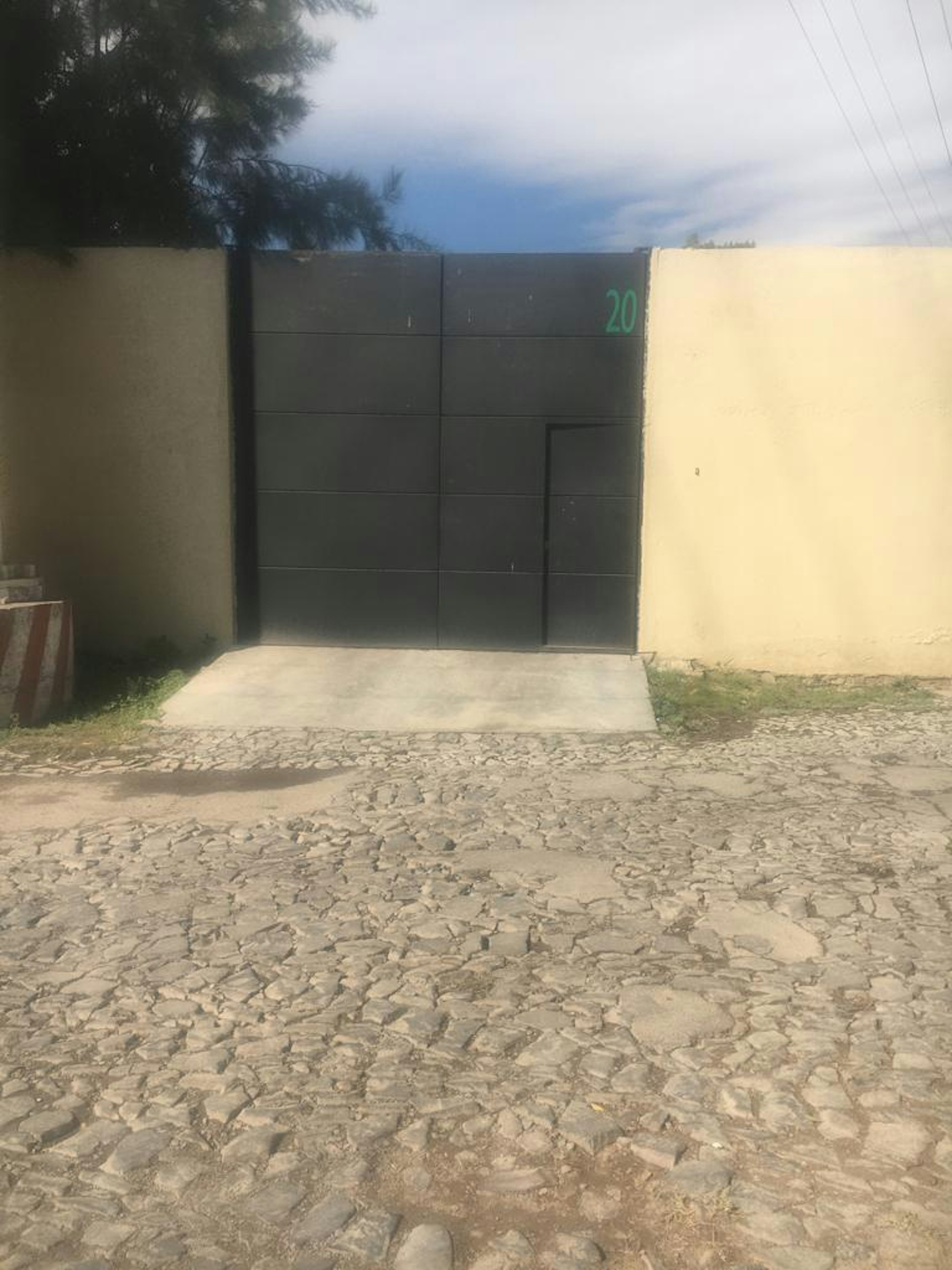 La imagen muestra la entrada de una propiedad con un portón metálico negro numerado 20. El portón está flanqueado por muros de color claro. Frente a la entrada hay un camino de piedras irregulares, típico de zonas residenciales tradicionales. El cielo azul y la vegetación visible sugieren un entorno suburbano o rural. La construcción parece sólida y bien mantenida, indicando una propiedad de calidad media a alta.