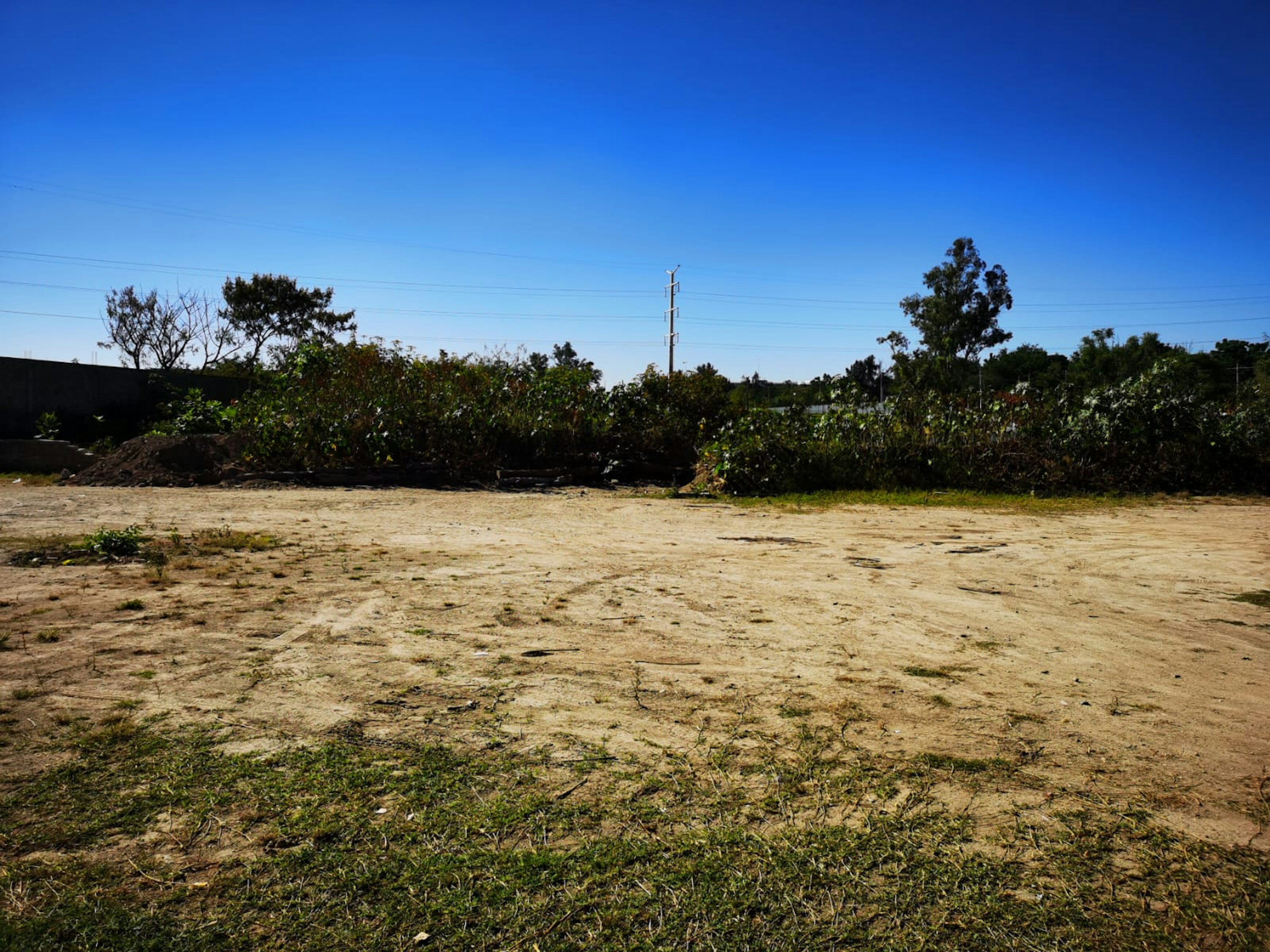 La imagen muestra un terreno baldío en una zona suburbana o rural. El suelo es arenoso y seco, con escasa vegetación. Al fondo se observan árboles y arbustos que delimitan la propiedad. Se ve una línea eléctrica que atraviesa el paisaje, sugiriendo que el área tiene acceso a servicios básicos. El cielo es de un azul intenso, indicando un clima seco. Este terreno podría ser ideal para desarrollo inmobiliario o agrícola.