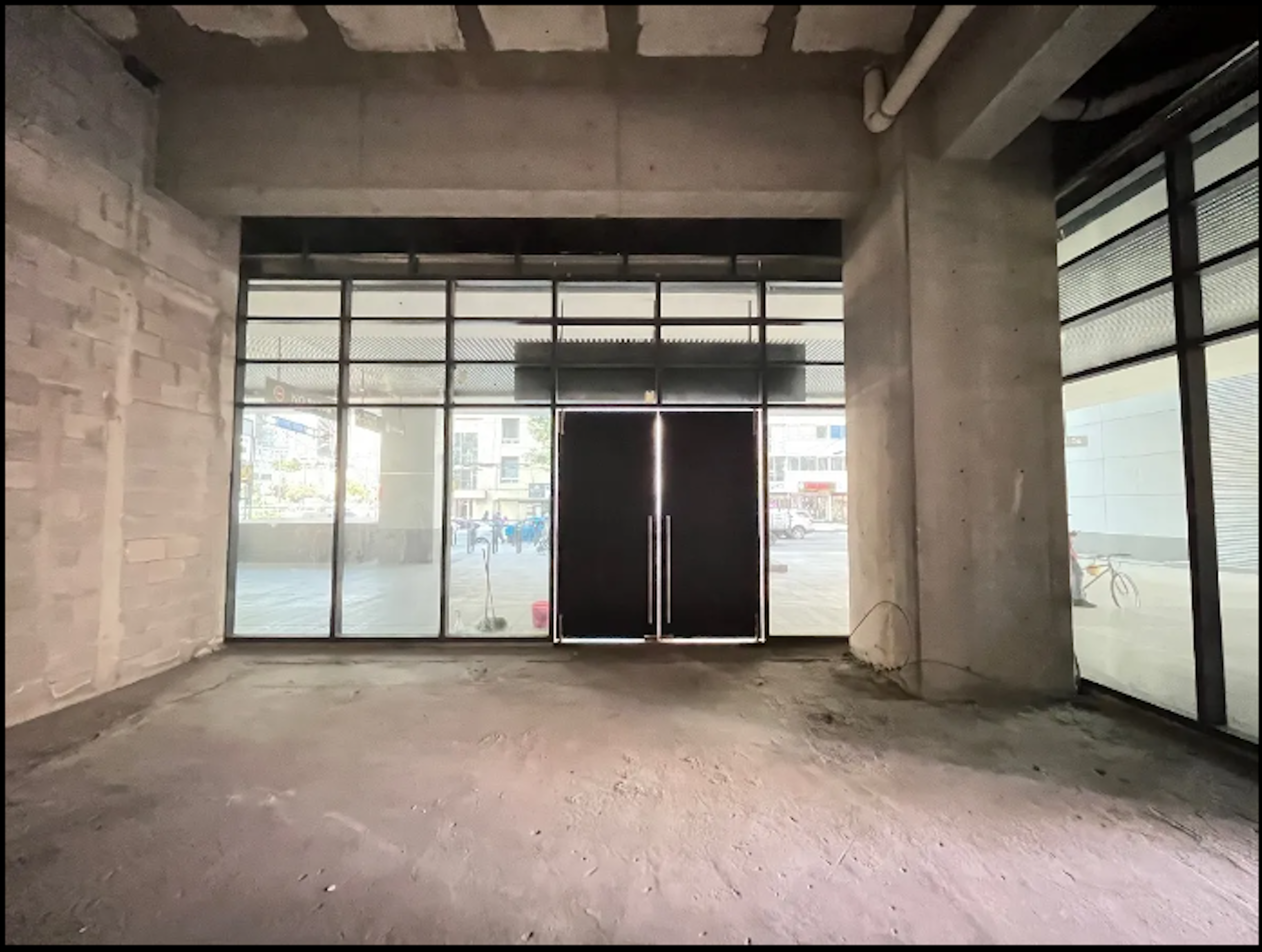 La imagen muestra un espacio comercial en construcción o remodelación. Se observa una entrada con puertas de vidrio y metal, rodeada de ventanales que dan a la calle. El interior tiene paredes de concreto expuesto y un piso sin acabados. El techo muestra vigas y tuberías visibles. El espacio parece estar en una etapa temprana de acondicionamiento, listo para ser personalizado según las necesidades del futuro inquilino.