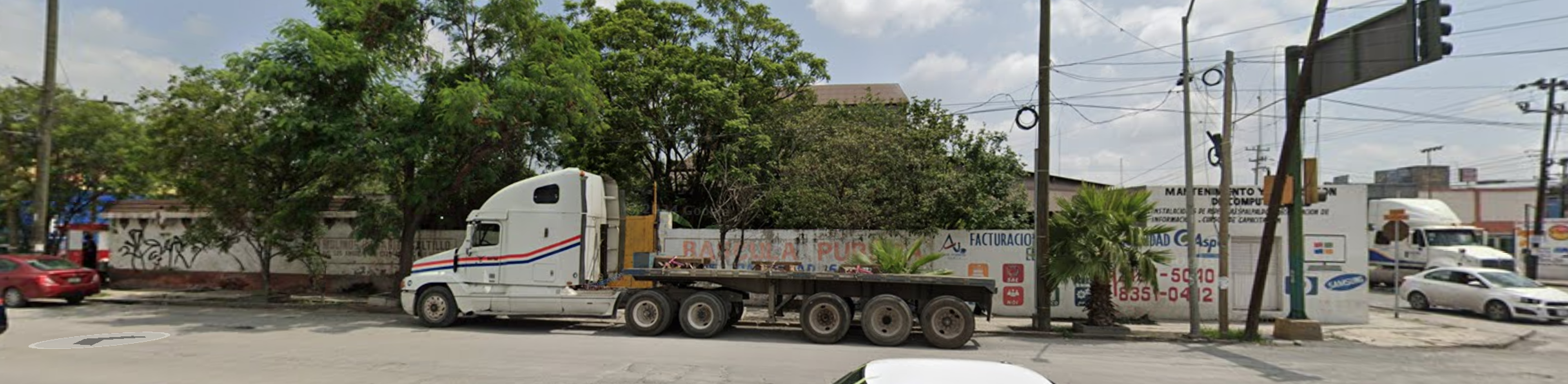 La imagen muestra una escena urbana con un gran camión de carga estacionado en una calle. El entorno tiene árboles, postes de electricidad y edificios bajos. Se observan algunos negocios y carteles publicitarios. La zona parece ser un área comercial o industrial, posiblemente en las afueras de una ciudad. El estado de la infraestructura y la presencia del camión sugieren actividad económica y potencial para desarrollo inmobiliario comercial.