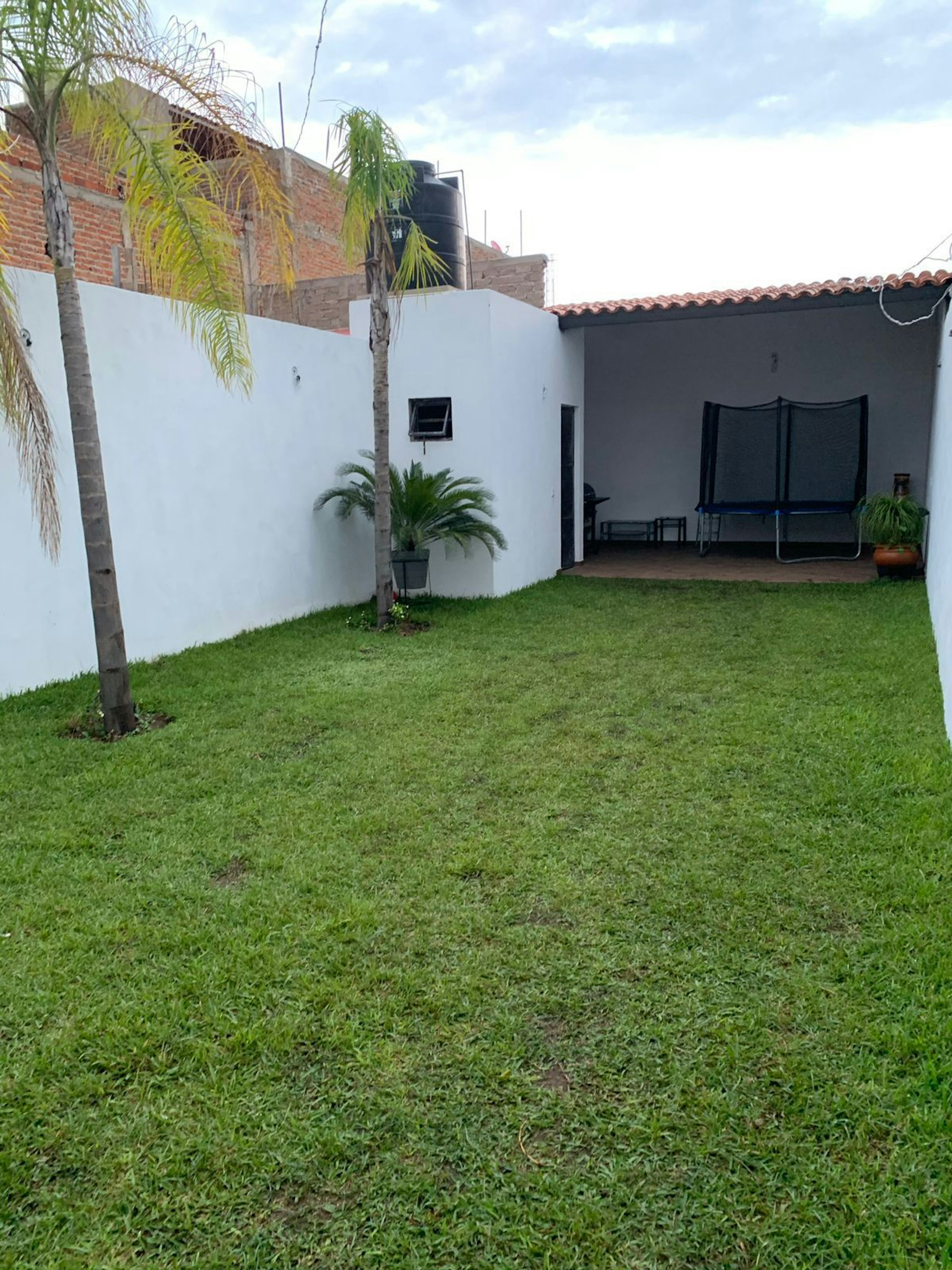 Espacioso jardín trasero con césped verde y bien mantenido, rodeado por muros blancos. Incluye una zona de patio con trampolín y mobiliario de exterior. Ideal para actividades al aire libre y reuniones familiares.