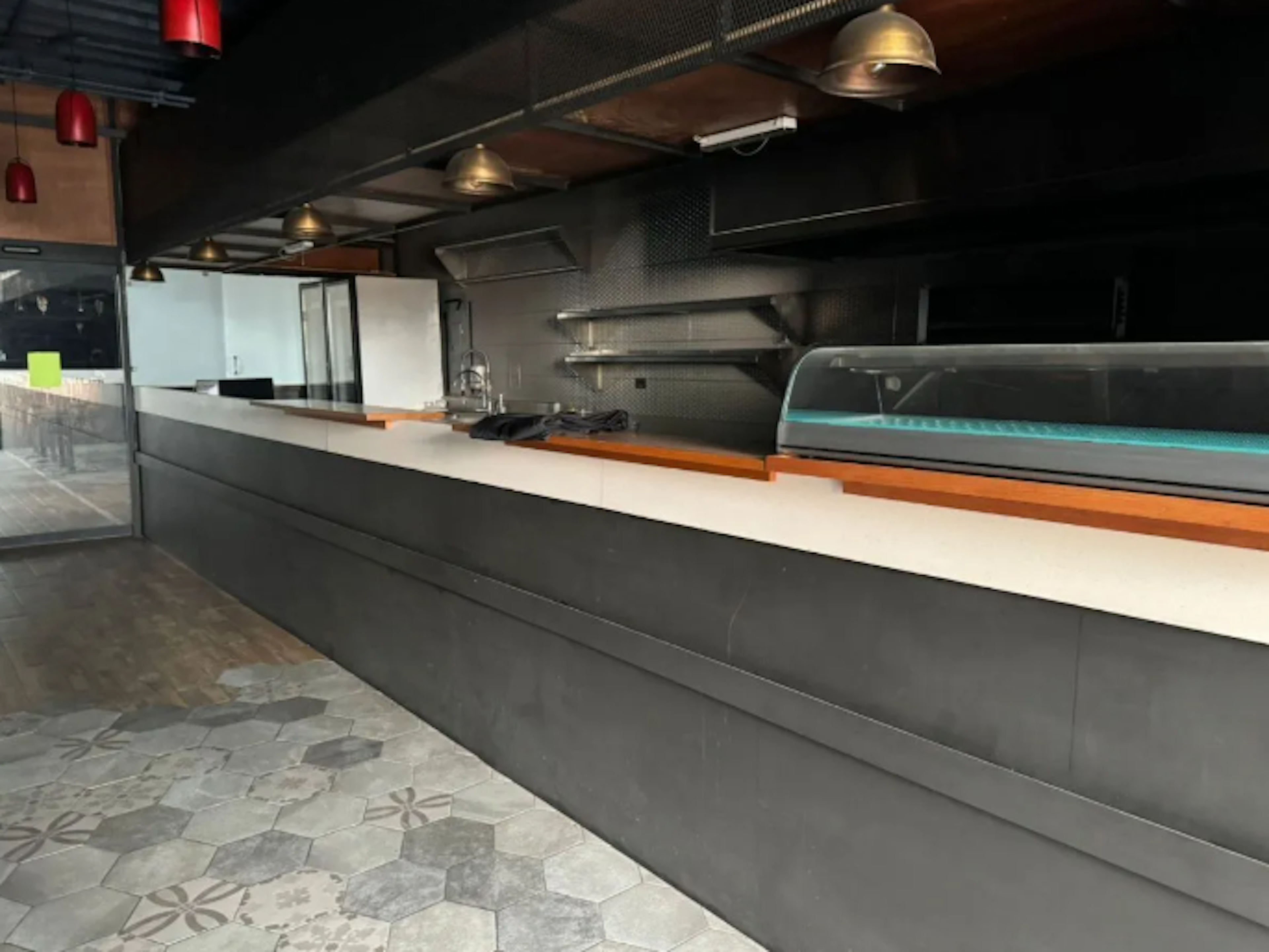 La imagen muestra el interior moderno de un establecimiento comercial, probablemente un restaurante o cafetería. Se aprecia un largo mostrador gris con una superficie blanca, equipado con una vitrina refrigerada. La cocina tiene acabados en acero inoxidable y estantes. El techo cuenta con lámparas colgantes rojas y doradas. El suelo tiene un diseño de baldosas hexagonales decorativas. El espacio tiene un estilo industrial y contemporáneo.