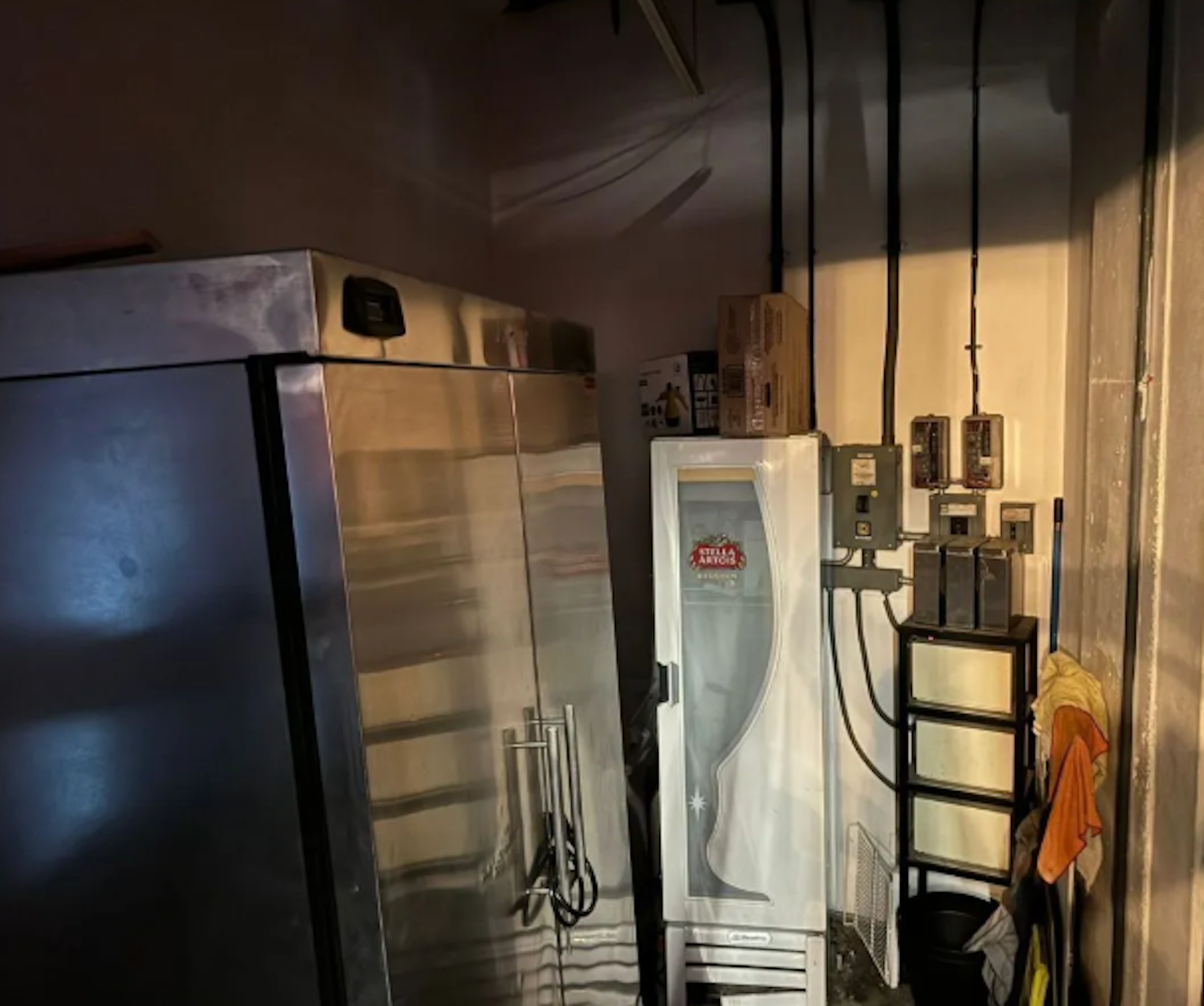 La imagen muestra un área de almacenamiento o cuarto de servicio con varios electrodomésticos y equipos. Se ve una nevera grande, un refrigerador de bebidas Stella Artois, paneles eléctricos y otros equipos industriales. El espacio parece estar en un sótano o área de servicio, con iluminación tenue y paredes sin acabados. La disposición sugiere un espacio utilitario en un edificio comercial o residencial multifamiliar.