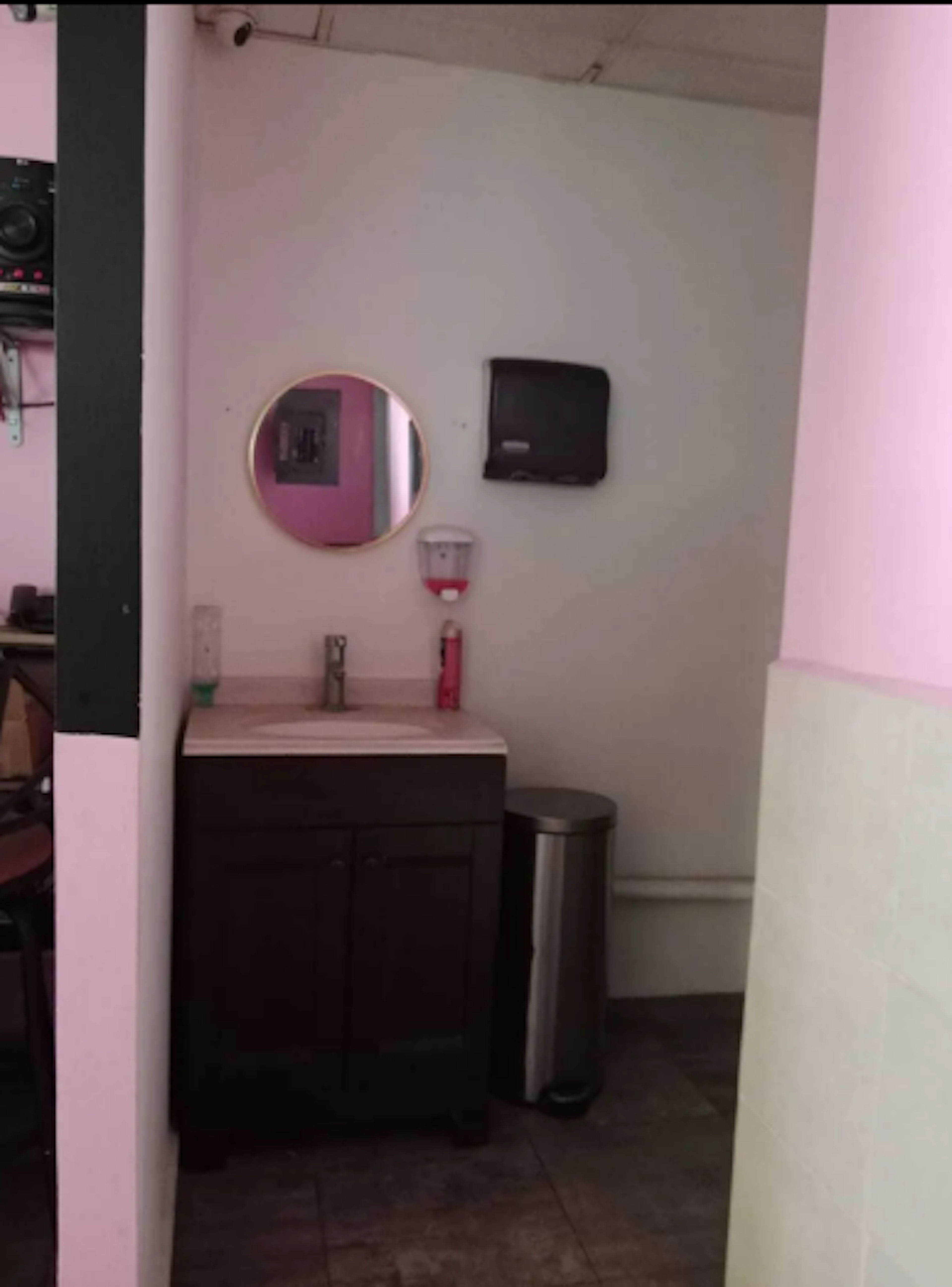 La imagen muestra un pequeño baño o área de aseo en un espacio comercial o de oficina. Tiene paredes rosa claro, un lavabo con gabinete oscuro, un espejo redondo, un dispensador de papel y un bote de basura. El diseño es minimalista y funcional, con accesorios básicos. La combinación de colores rosa y marrón oscuro da un toque moderno al espacio, que parece estar en buenas condiciones y bien mantenido.