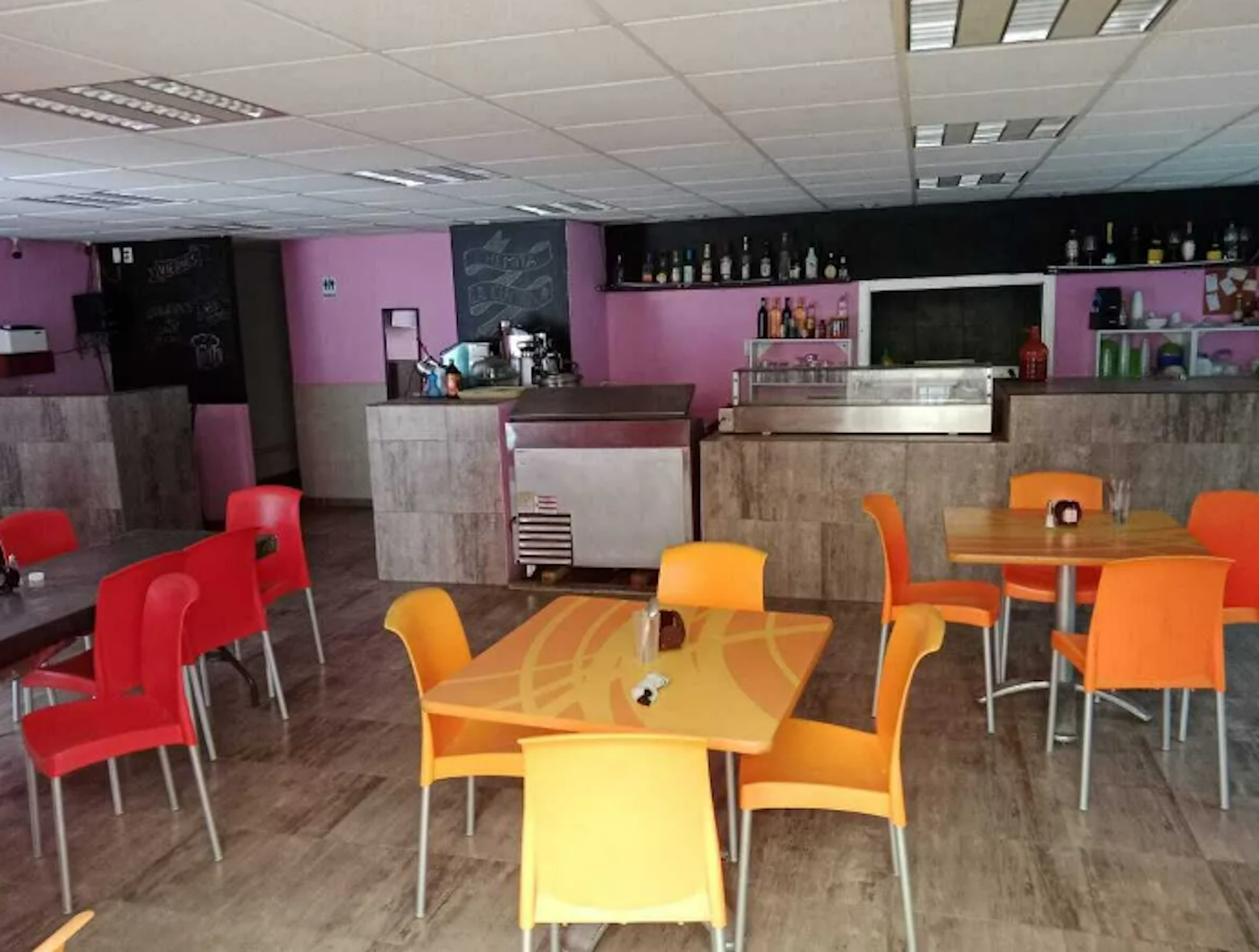 La imagen muestra el interior de un bar o cafetería con paredes rosas y una barra de servicio. El espacio tiene mesas y sillas coloridas en amarillo y rojo. Se observa una zona de bar con botellas de licor en estantes. El local tiene un diseño moderno y acogedor, con iluminación de techo y suelo de madera, ideal para un negocio de hostelería o restauración.