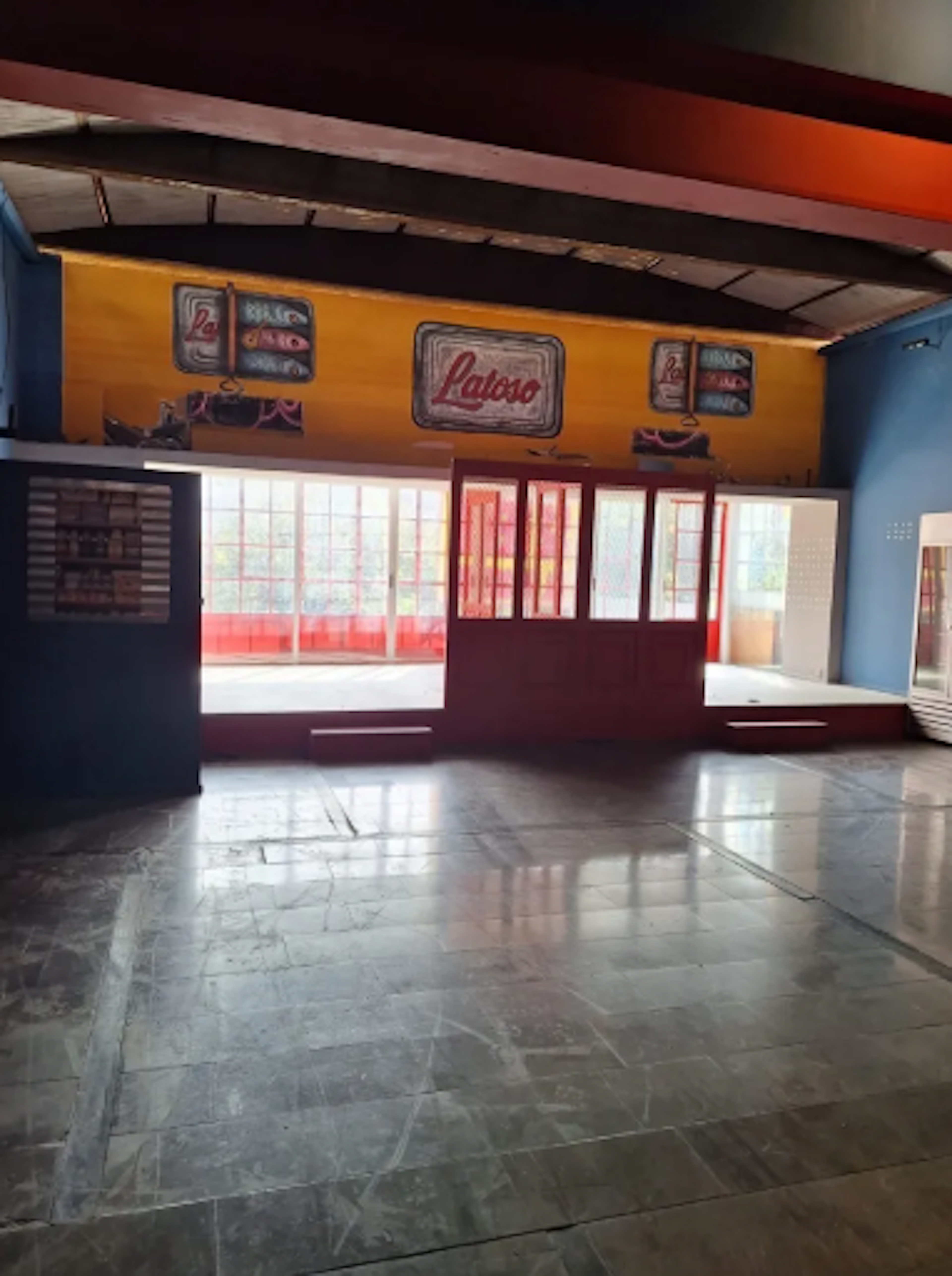 La imagen muestra el interior de un local comercial o espacio de venta, posiblemente una tienda o un quiosco. Tiene una pared amarilla con logos de Laioso, ventanas grandes con marcos rojos, y un suelo de baldosas brillantes. El espacio parece vacío y listo para ser ocupado o remodelado. Los colores vibrantes y el diseño sugieren un ambiente retro o vintage.