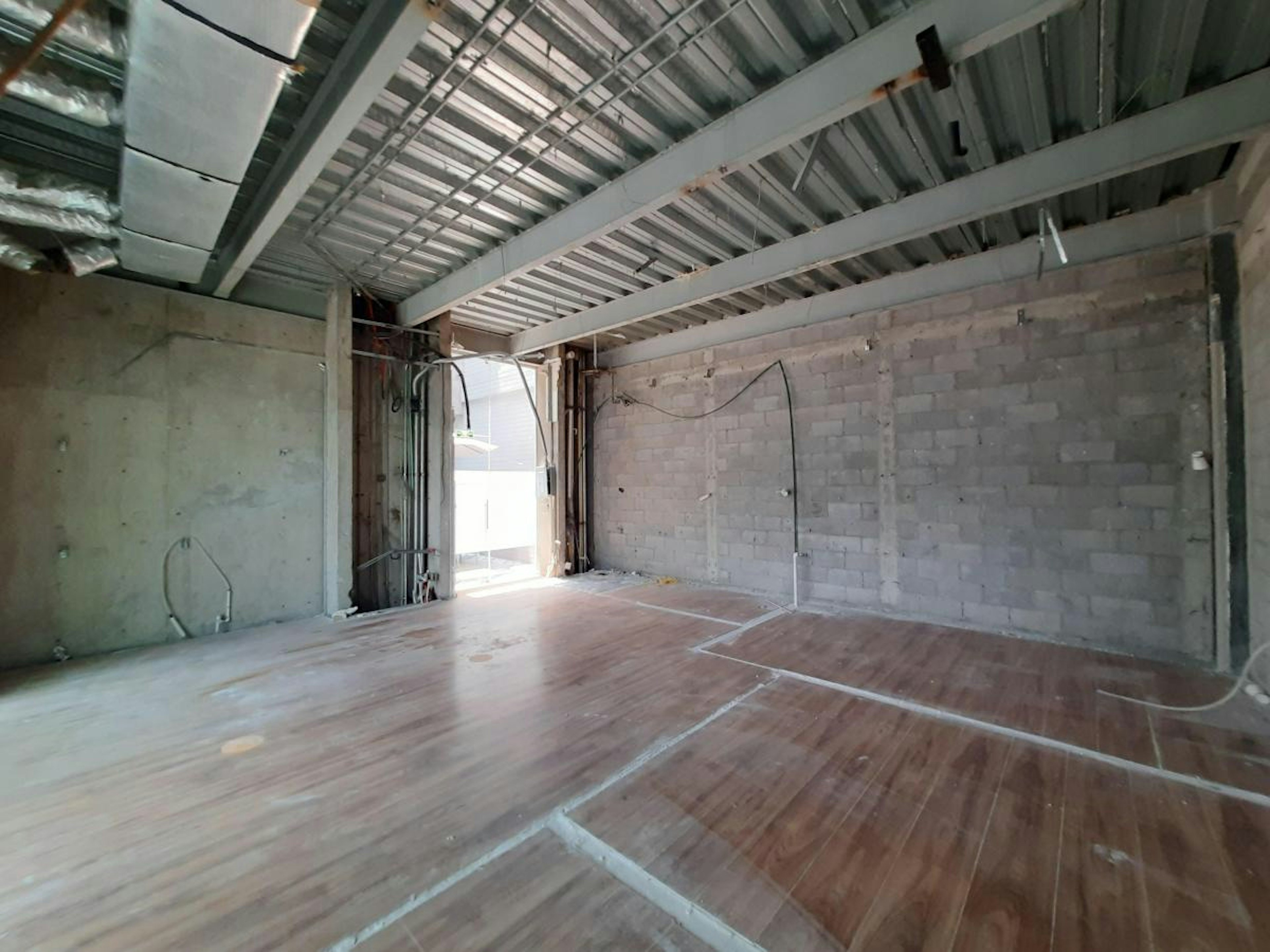 La imagen muestra un espacio interior en construcción o renovación. Se observa una estructura metálica en el techo, paredes de ladrillo y concreto sin acabados, y un piso de madera parcialmente instalado. Hay una abertura que podría ser una puerta o ventana, permitiendo la entrada de luz natural. El espacio parece amplio y diáfano, ideal para una futura oficina, loft o área comercial en desarrollo.